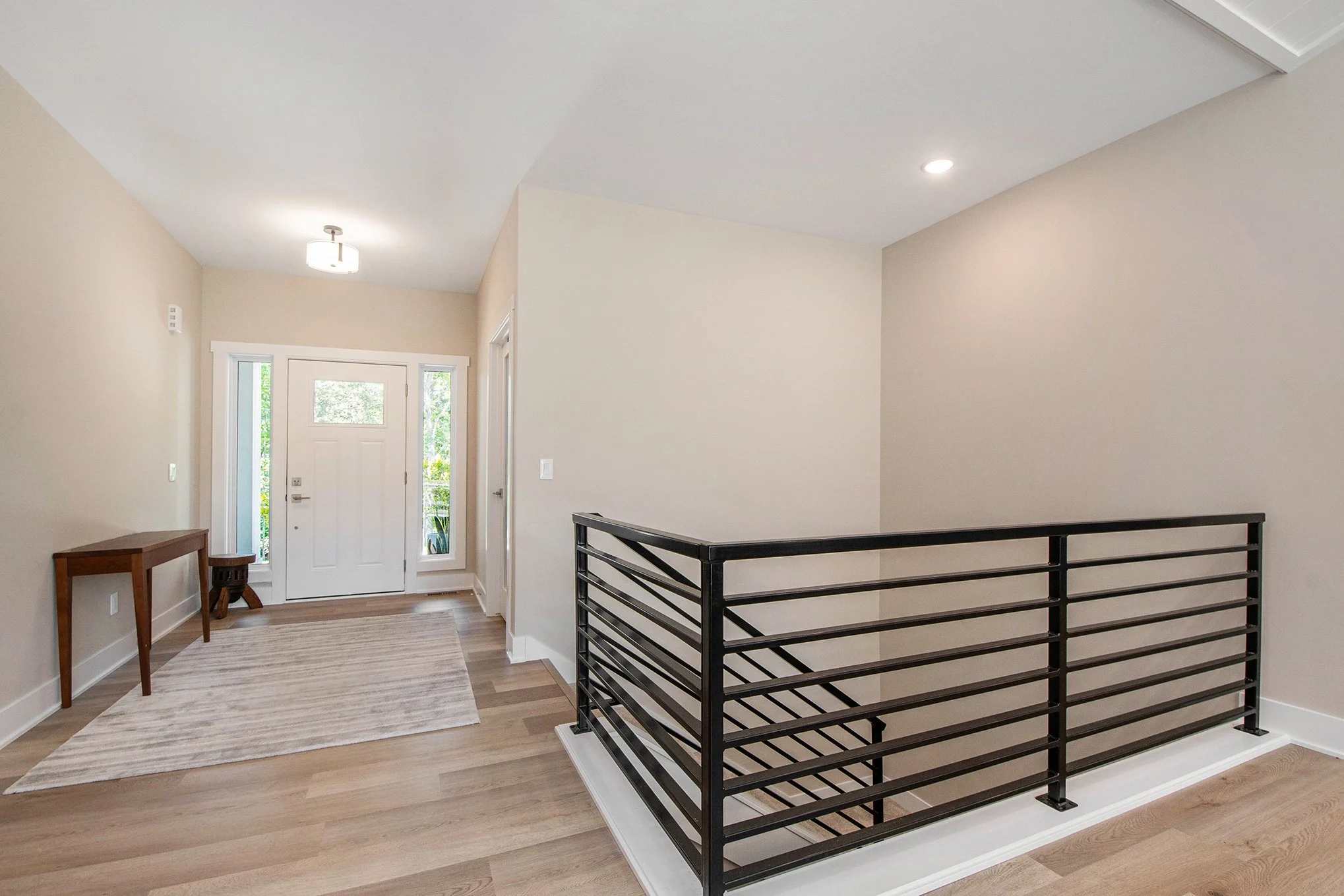 Bright entryway and staircase with black metal railing, light-colored walls, wooden floor, front door with side windows, small wooden table