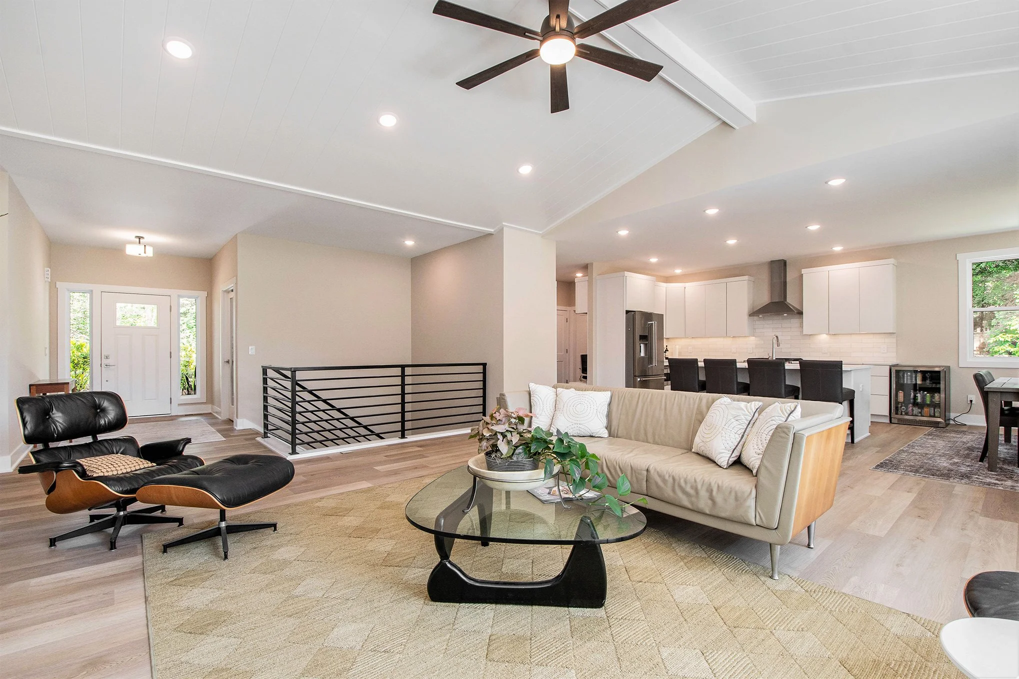 Living room with black lounge chair, beige sectional sofa, glass coffee table, plants, open kitchen with white cabinets, black chairs, stainless steel appliances, ceiling fan, and hardwood floors.