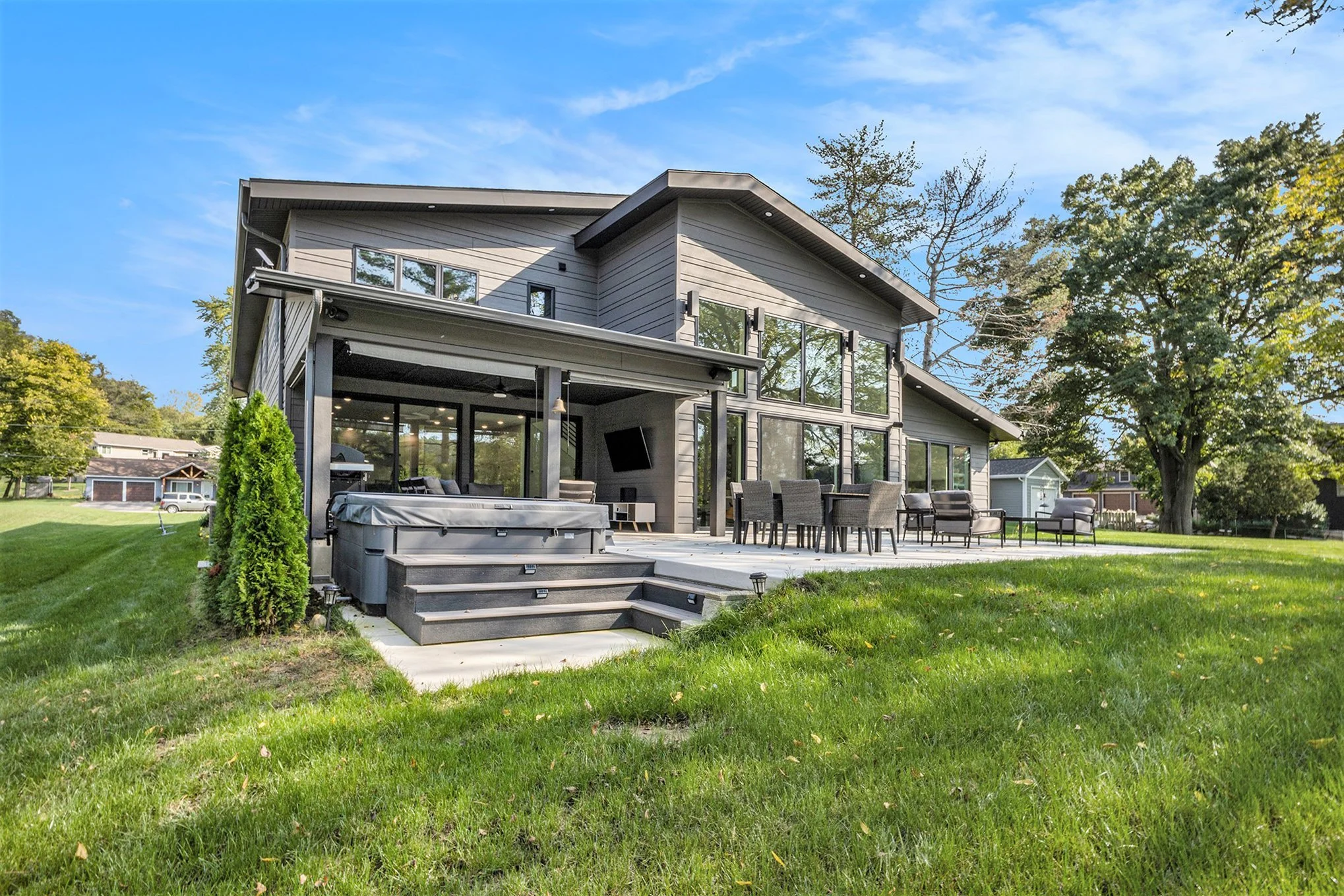 Modern two-story house with large windows, outdoor patio with seating, hot tub, surrounded by green grass and trees, under a blue sky.