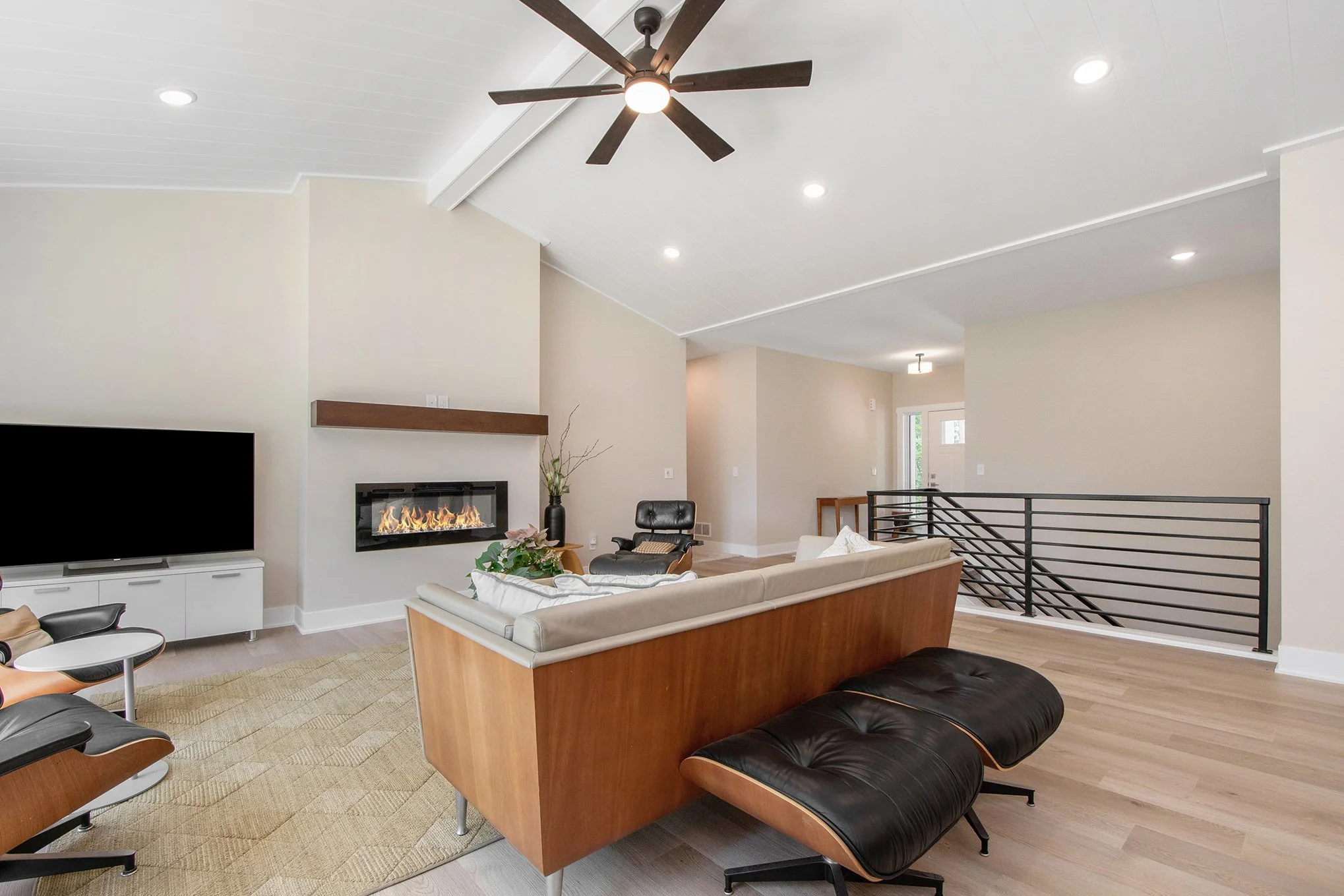Modern living room with a black ceiling fan, recessed lighting, a fireplace, a large flat-screen TV, beige walls, and wooden furniture.