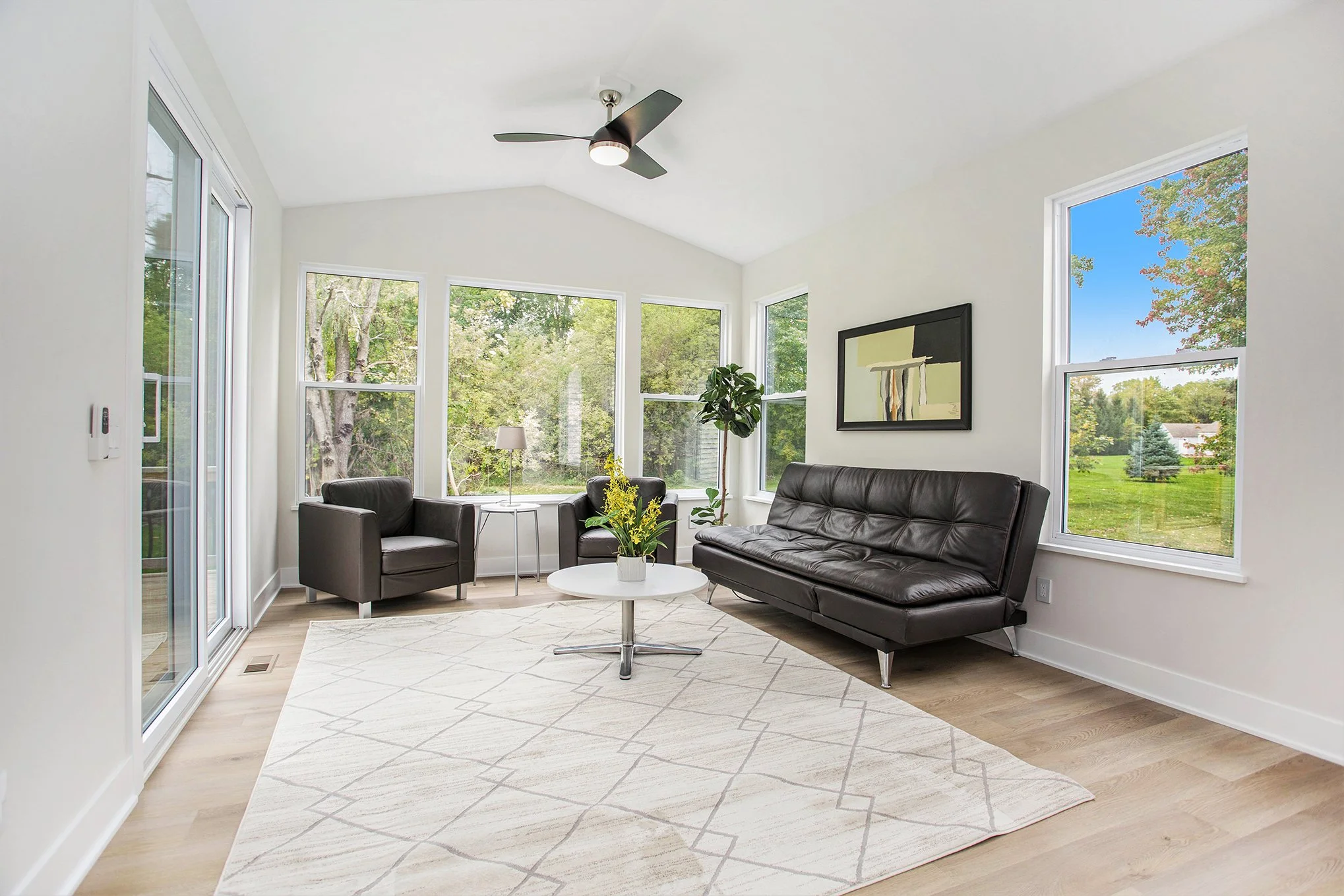 Bright living room with white walls, large windows overlooking greenery, black leather sofa, black armchairs, a round white coffee table, a floor lamp, and abstract wall art.