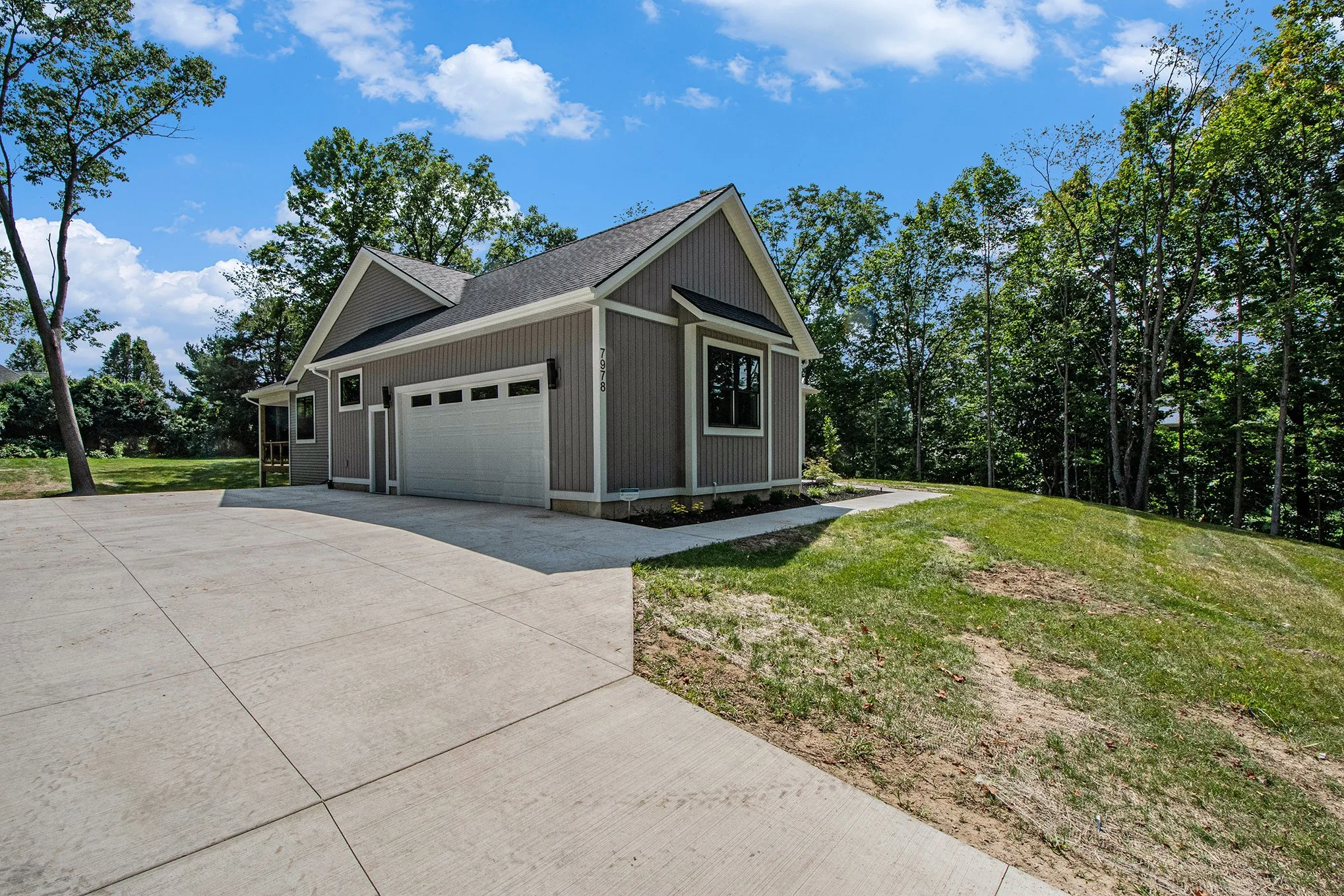 Newly built gray house with a white garage door, surrounded by green trees and a grassy lawn on a sunny day with blue sky and clouds.