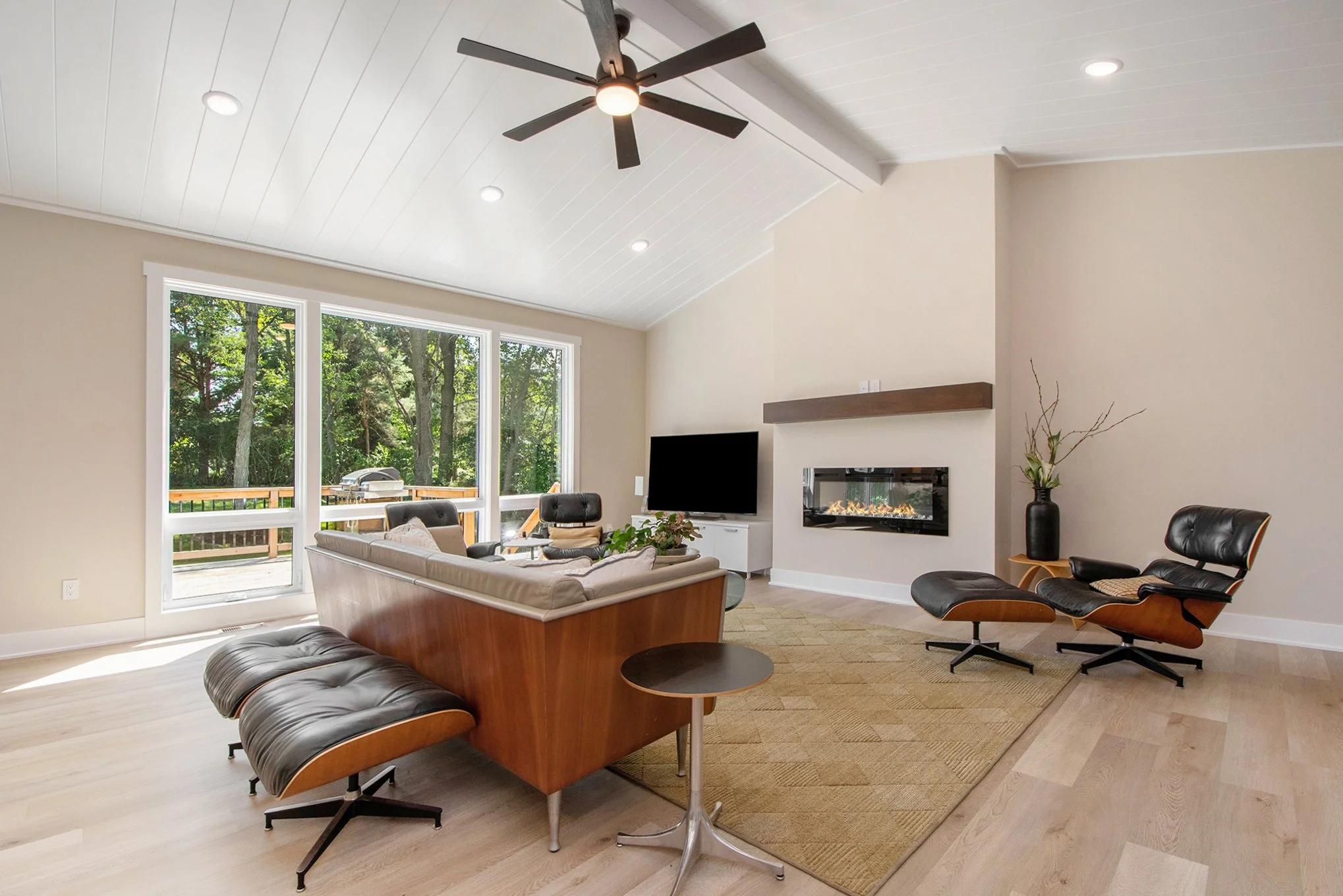 Modern living room with large window, ceiling fan, leather armchair with ottoman, sofa, wall-mounted TV, fireplace, and decorative plant.