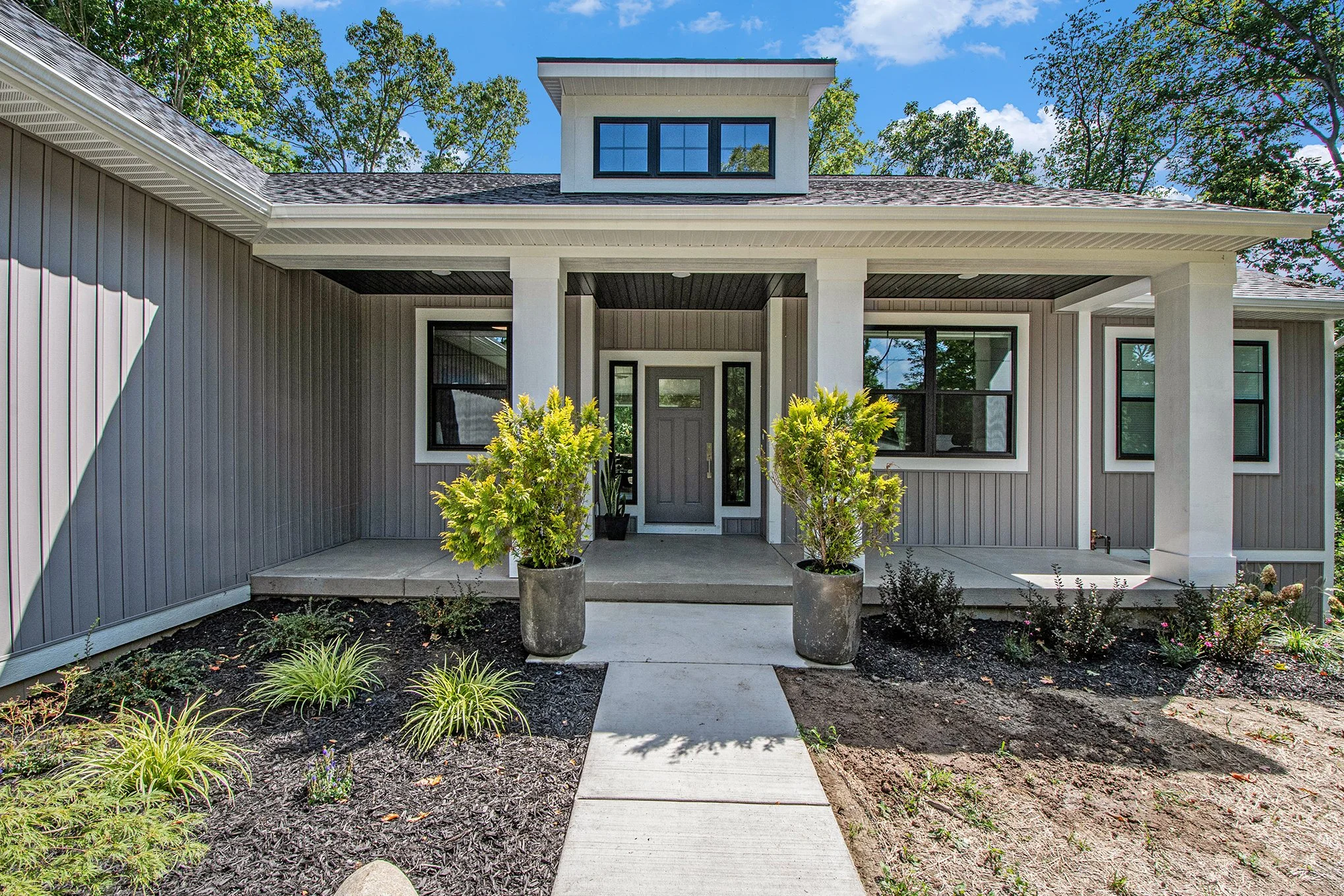 Front view of a modern house with a porch, gray siding, black window frames, and a small dormer on the roof. Potted plants are on the porch, and the yard has some plants and soil under a clear blue sky.