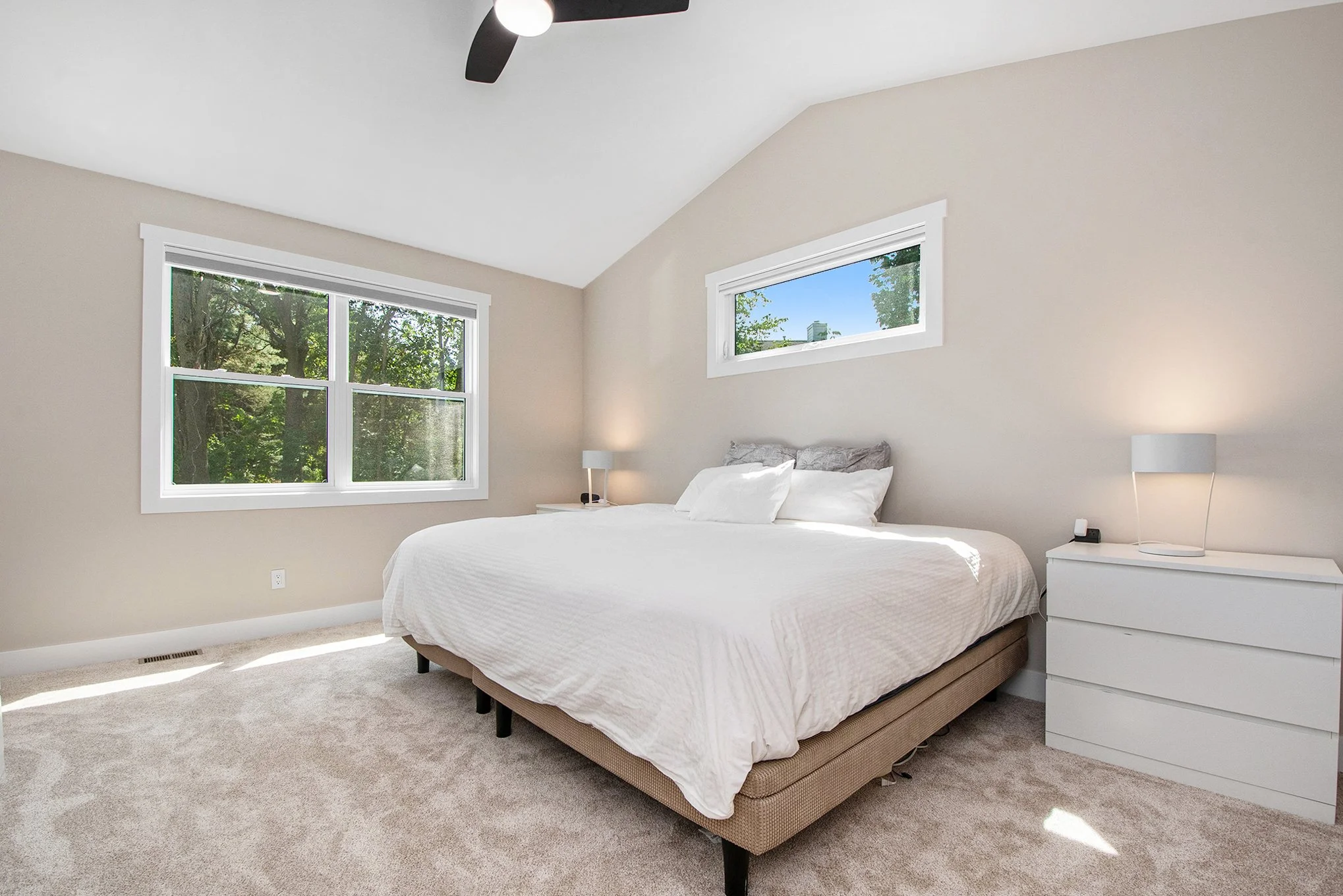 A bedroom with beige walls, a large window with trees outside, a white bed with pillows, two white nightstands with lamps, a ceiling fan, and a broad window on the wall.