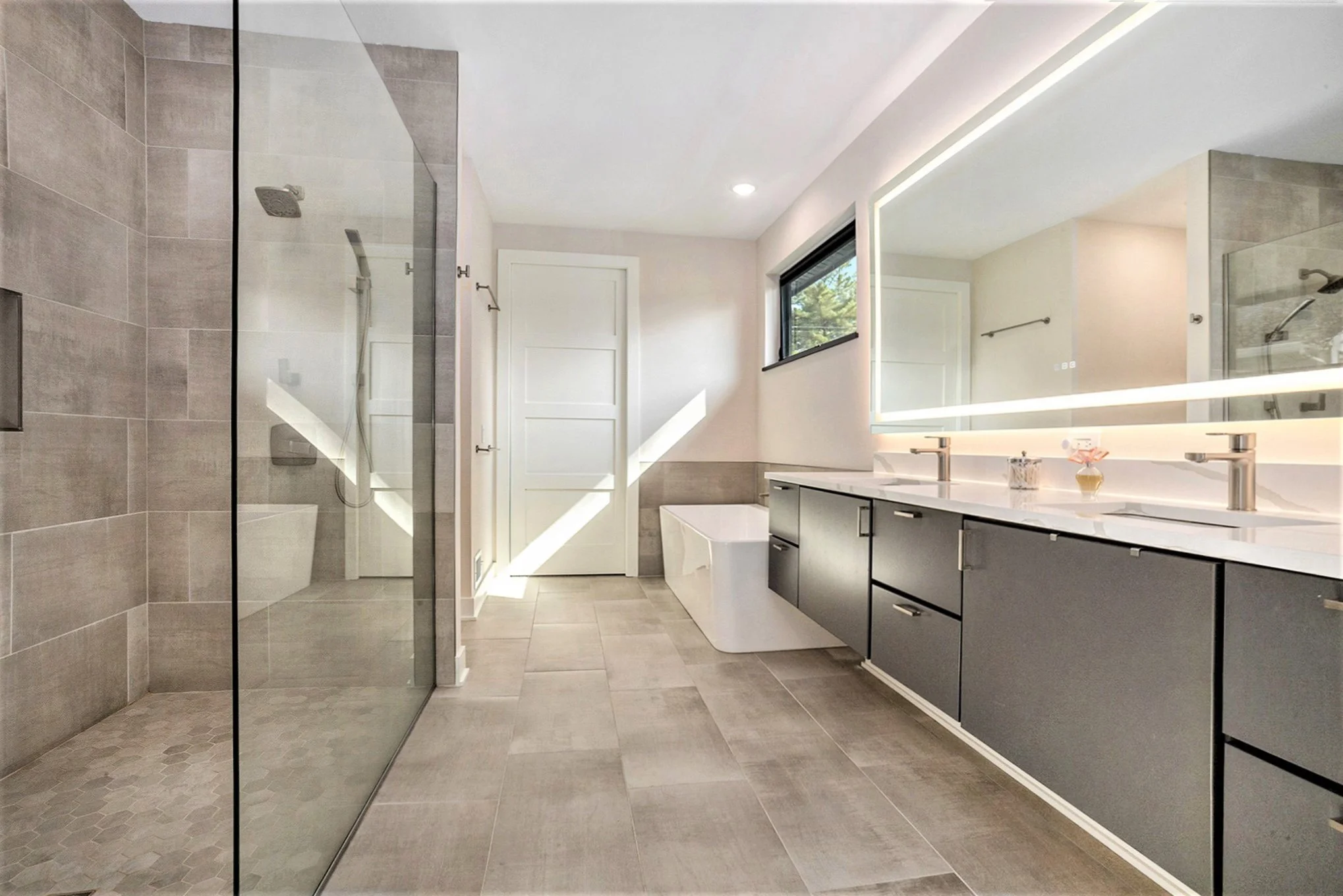 Modern bathroom with large walk-in shower with glass door, double vanity with large mirror and lighting, small window, and a bathtub in the corner. Neutral-colored tiles and cabinetry.