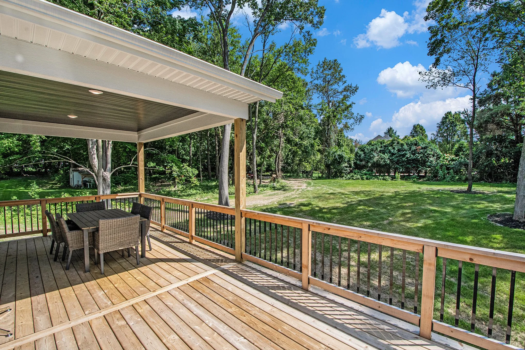 Wooden deck with outdoor dining table and chairs overlooking a green yard with trees and a blue sky with fluffy clouds.