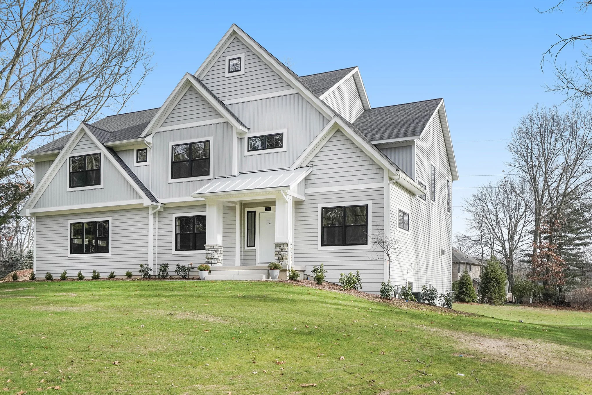 A large white modern house with multiple gabled roofs, black window frames, and a small front porch with two pillars, situated on a lawn with trees around.