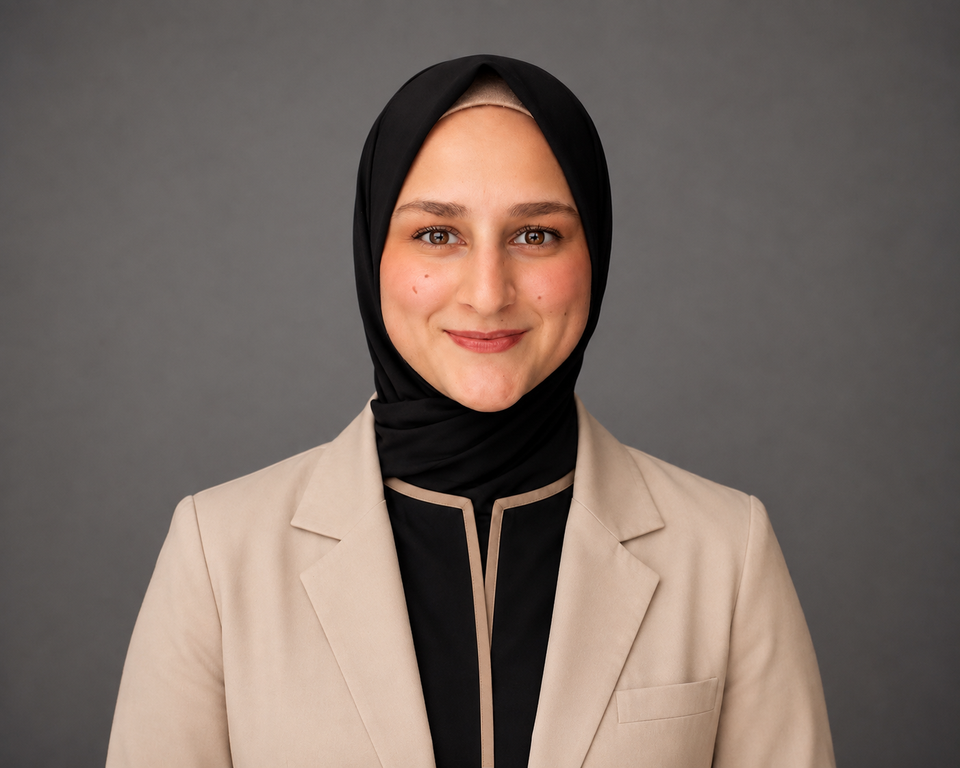 A young woman in a black hijab and graduation gown standing on outdoor steps with a stone building in the background.