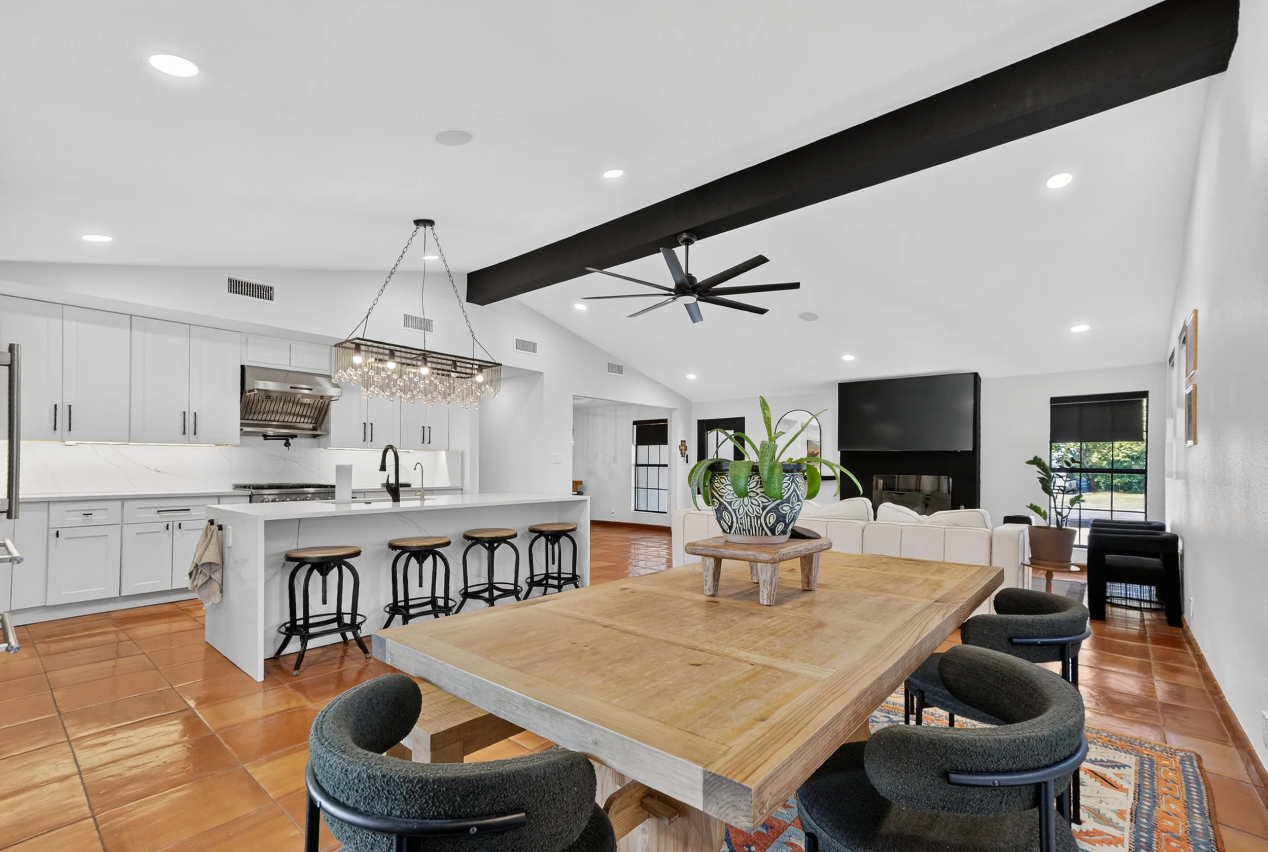 Open-concept kitchen and living room with white cabinets, a large wooden dining table, black chairs, a chandelier, a ceiling fan, and large windows with blinds.