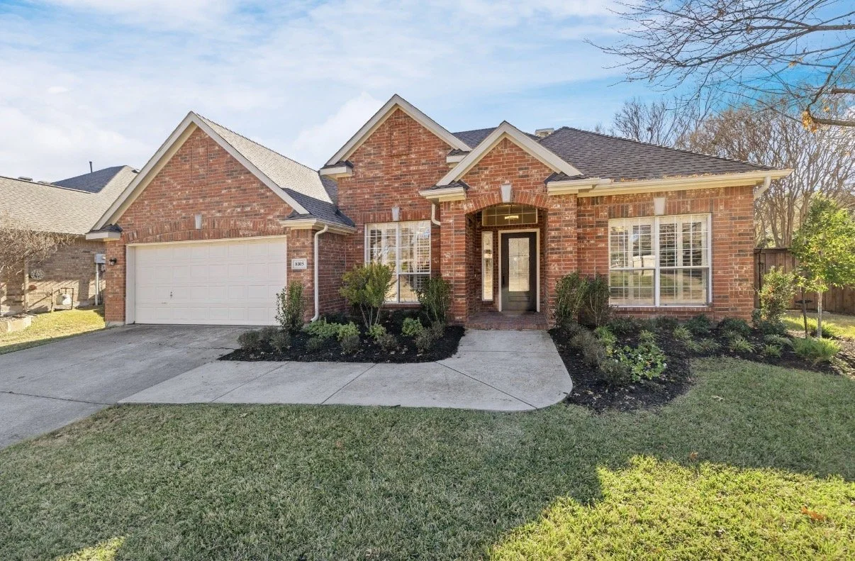 A red brick single-family house with a white garage door, front lawn, and landscaped garden with small bushes and mulch.