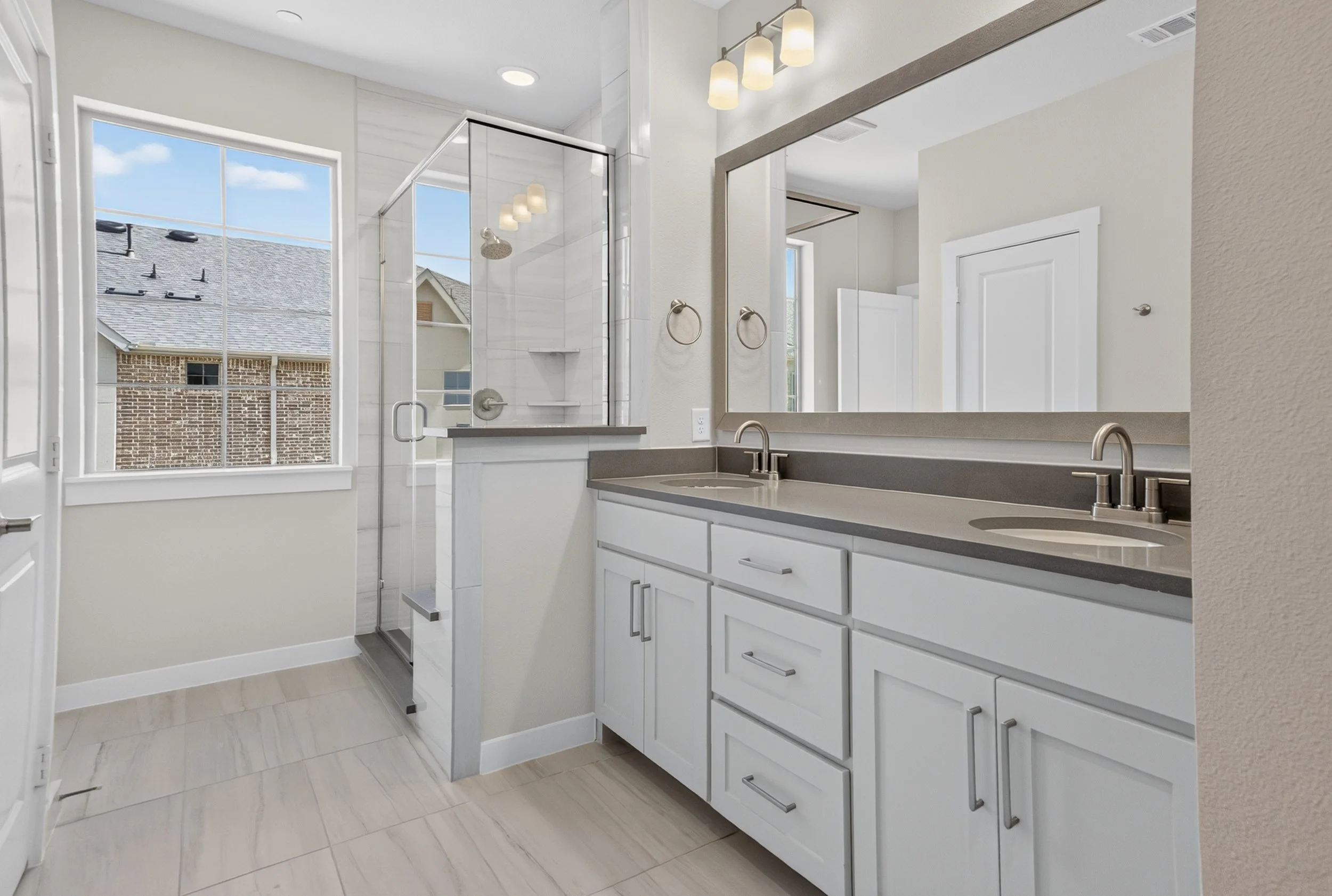 Modern bathroom with double sinks, white cabinetry, large mirror, and natural light from a window with a view of neighboring rooftops.