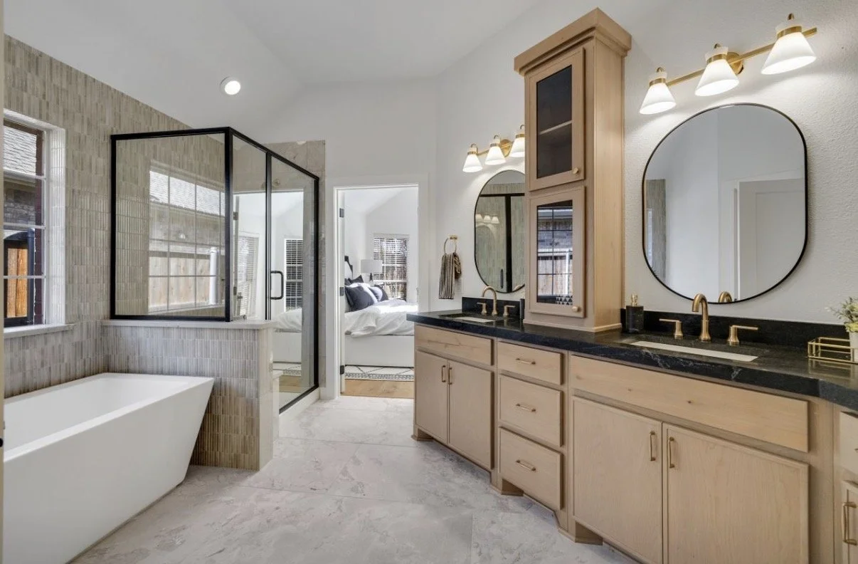 Bathroom with dual vanities, large mirrors, gold fixtures, a bathtub, and a separate shower area, connected to a bedroom with a bed and windows.