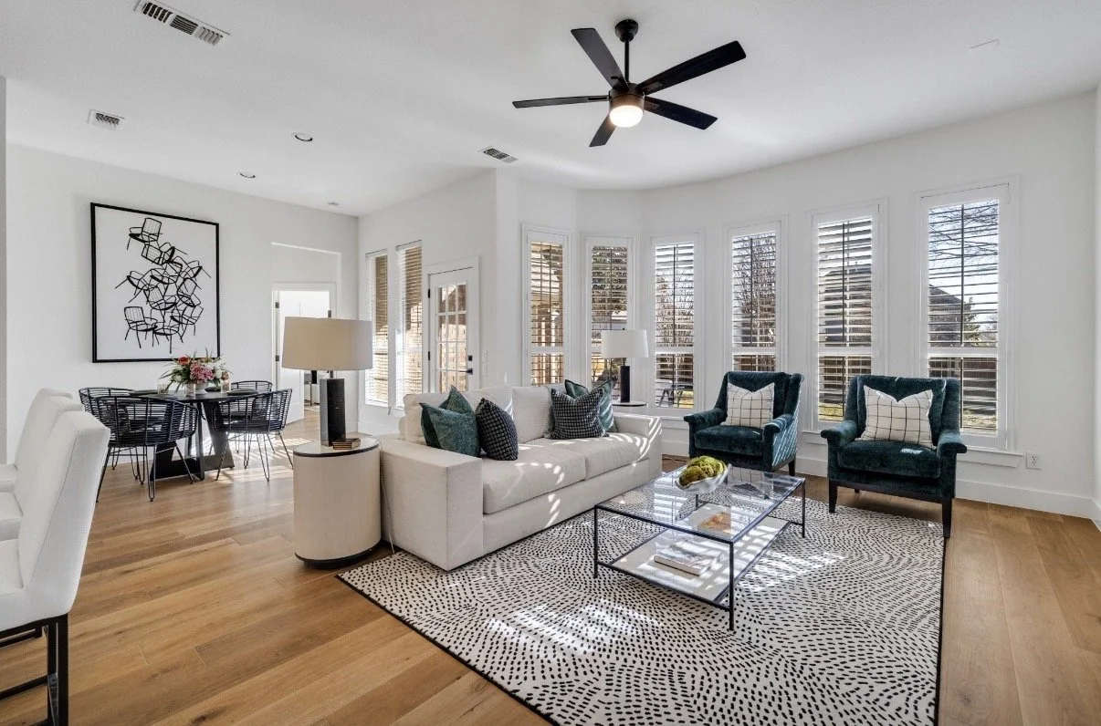 Living room with white walls, large windows with blinds, a ceiling fan, a white sofa with multiple pillows, a glass coffee table, two dark green armchairs with pillows, a patterned rug, and a black and white wall art piece near a dining area.