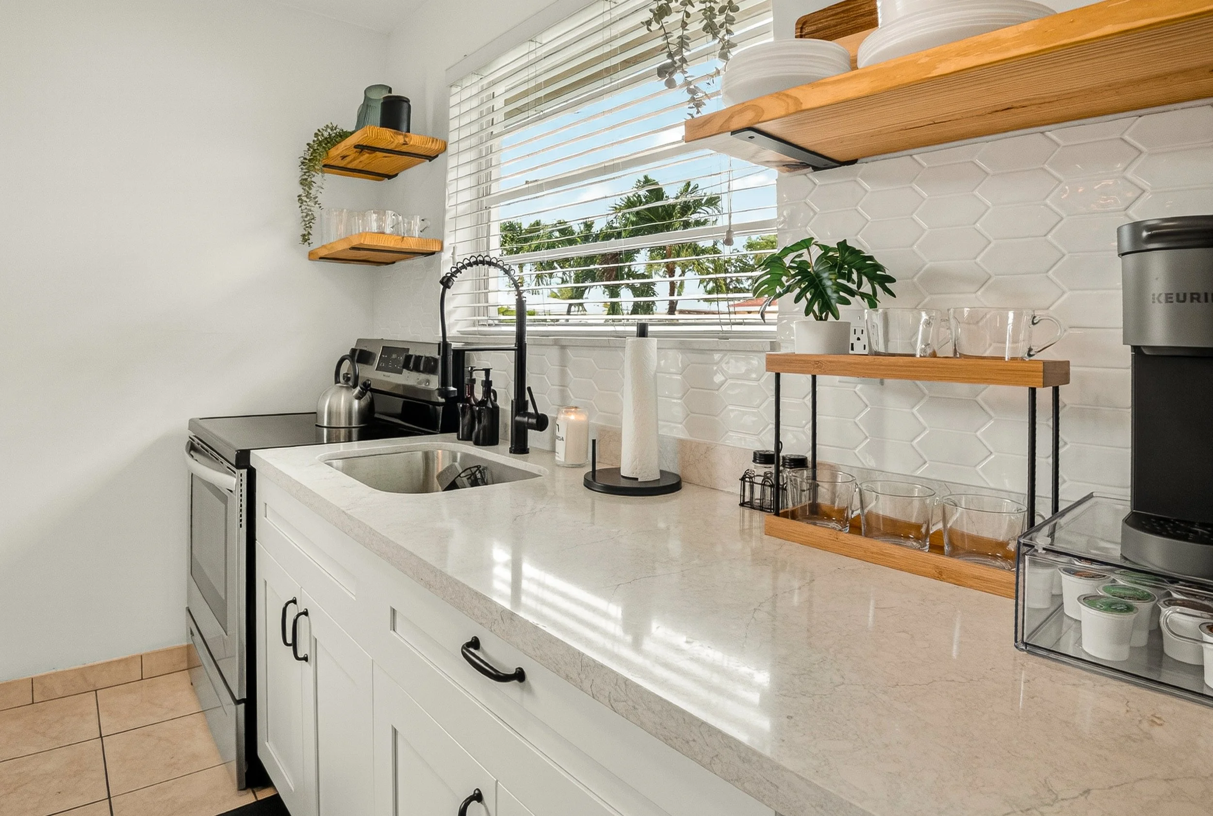 Kitchen with white cabinets, a marble countertop, a window with blinds showing palm trees outside, open wooden shelves with dishes and decor, a sink with a gooseneck faucet, a paper towel holder, a coffee maker, and glassware on a nearby shelf.
