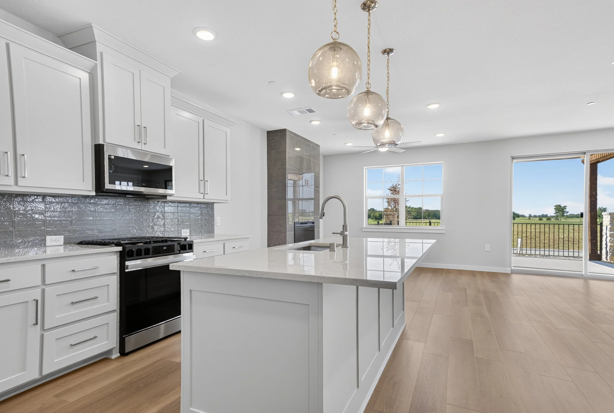 Modern kitchen with white cabinets, stainless steel appliances, a large island, pendant lights, and windows overlooking a rural landscape.