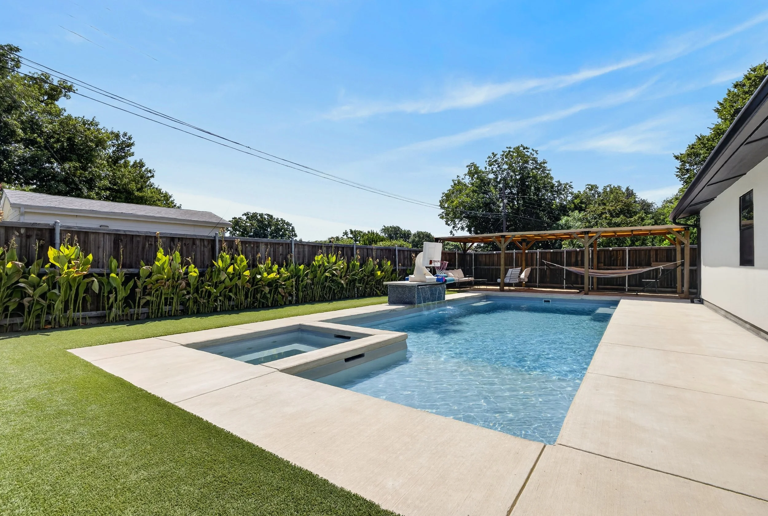 Backyard with a swimming pool, hot tub, green lawn, wooden fence, trees, a house to the right, and a shed or additional building to the left. Clear blue sky with a few clouds.
