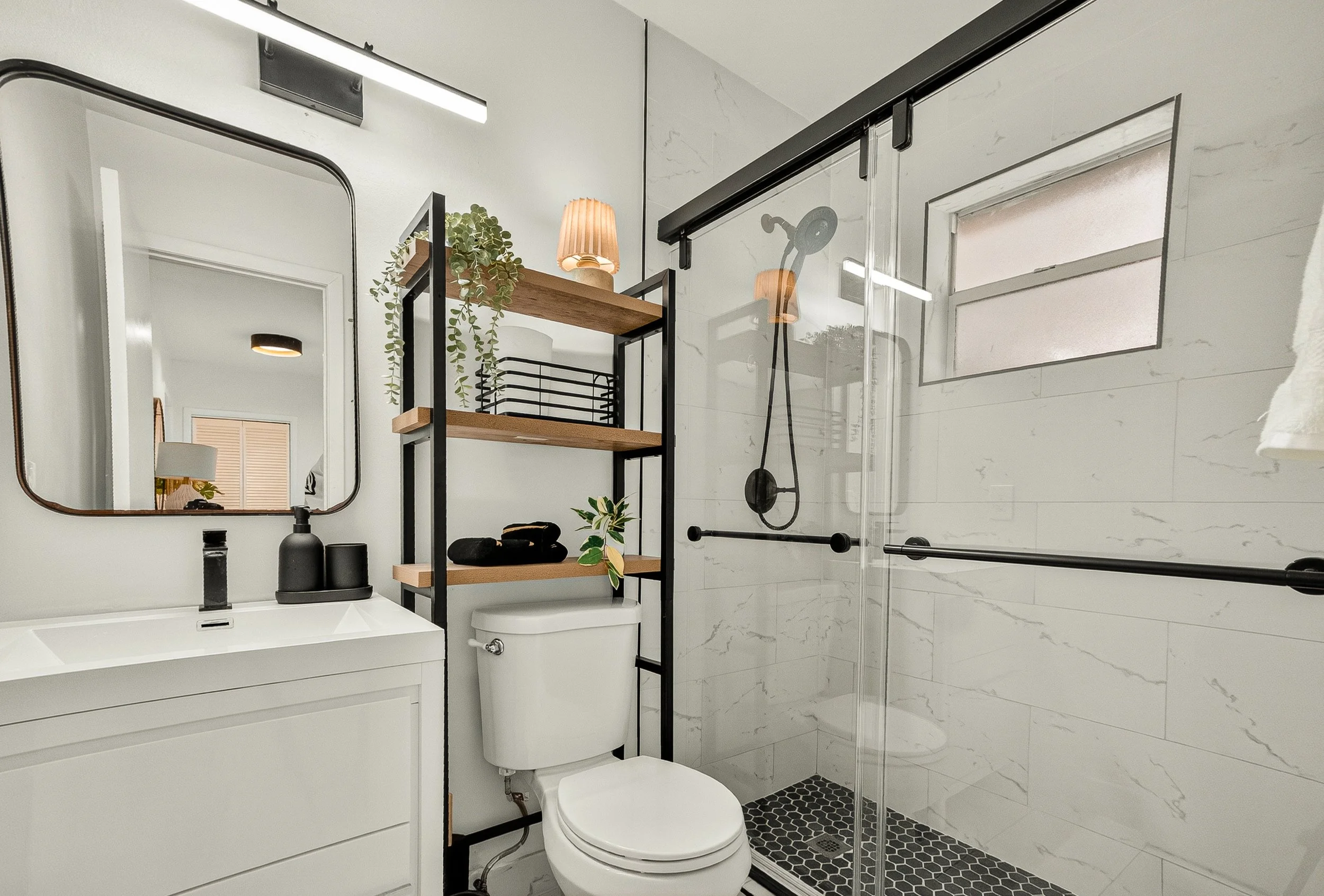 Modern bathroom with white vanity, mirror, black fixtures, shelving with plants and decor, toilet, and glass enclosed shower with white marble walls and black grid tile floor.