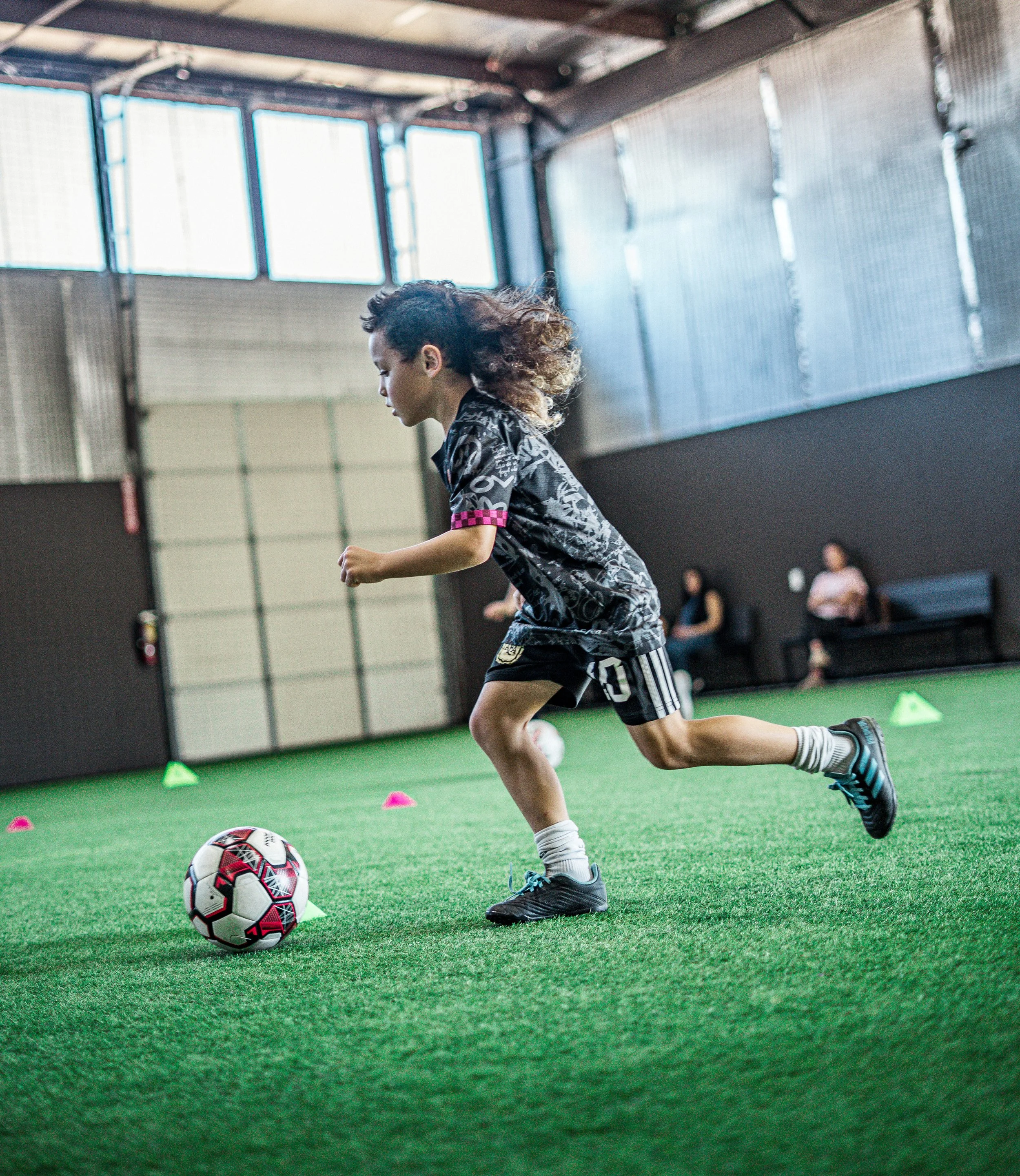 A young girl playing soccer indoors on a green turf field, wearing a patterned sports jersey, black shorts, and black soccer shoes, dribbling a soccer ball.