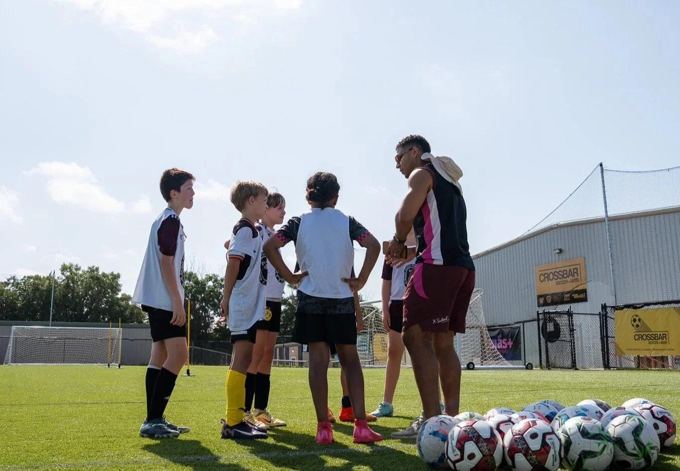 A group of young soccer players standing on a field in a circle listening to their coach during practice, with soccer balls on the grass nearby.
