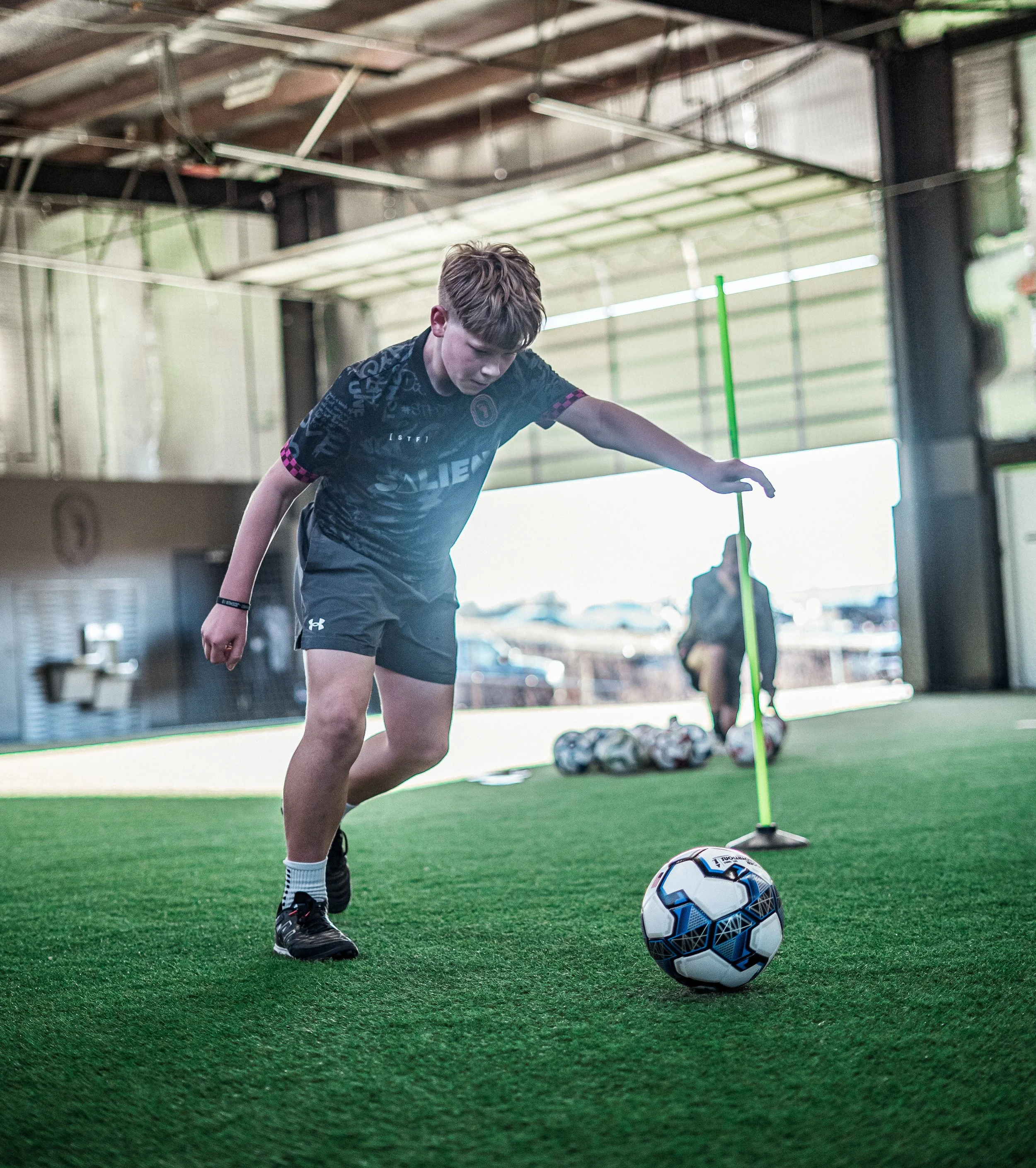 A young boy in sports attire practicing soccer indoors. He is focused on a soccer ball in front of him on a turf field with equipment and a goal in the background.