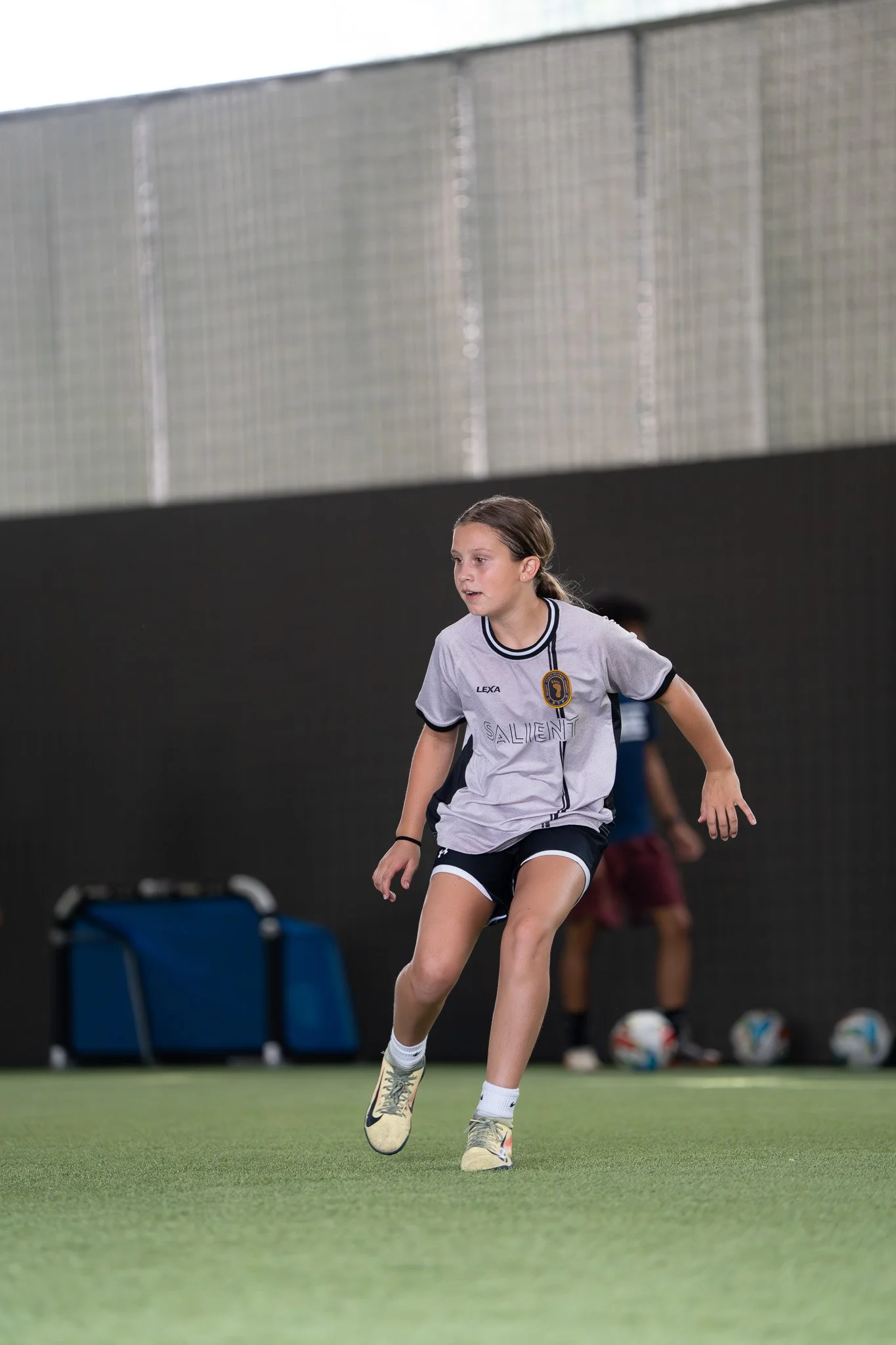 A young female soccer player wearing a gray and black uniform with the word 'Salient' on it is running on an indoor field with artificial turf.