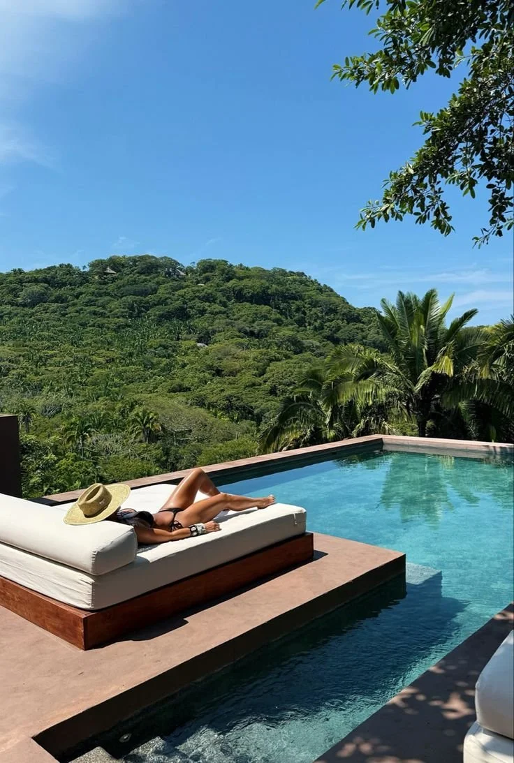 A woman relaxing on a poolside lounge chair with a straw hat, overlooking lush green hills under a bright blue sky.