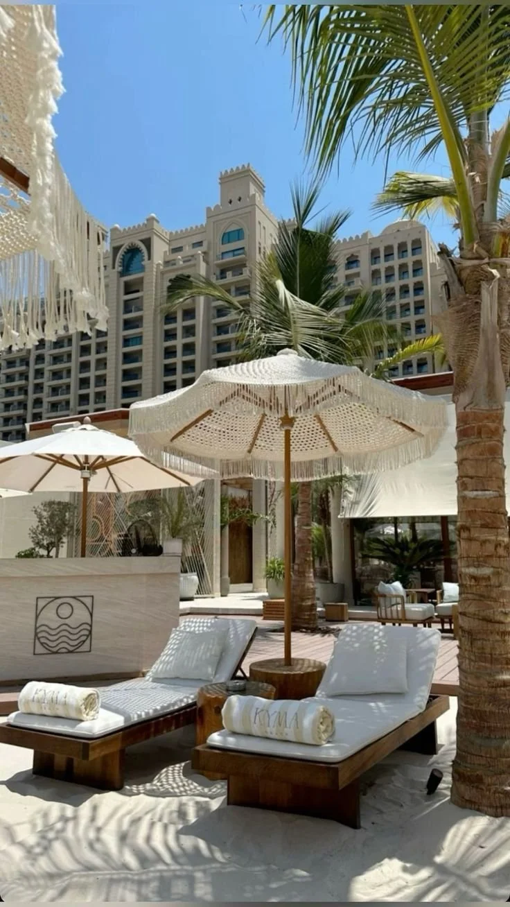 Beachside lounge chairs with umbrellas and towels in front of a hotel with palm trees and a clear blue sky.
