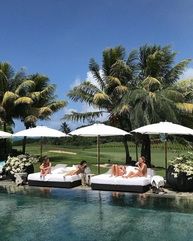 Four women relaxing on poolside beds under white umbrellas, with palm trees and a clear blue sky in the background.