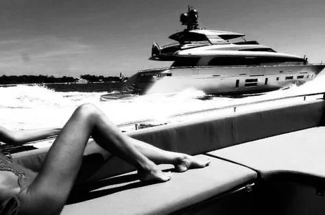 A person reclining on a boat with a large yacht in the background, in a black and white photo.