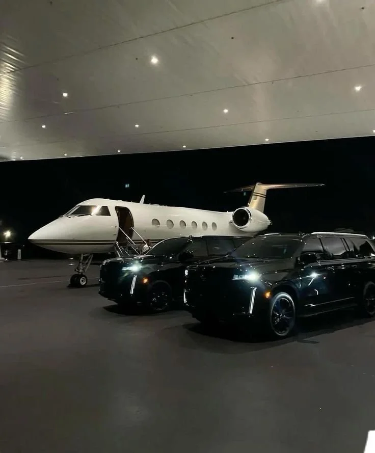 Nighttime view of two black luxury cars parked in front of a private jet on a tarmac, under a large, curved hangar roof with small lights.