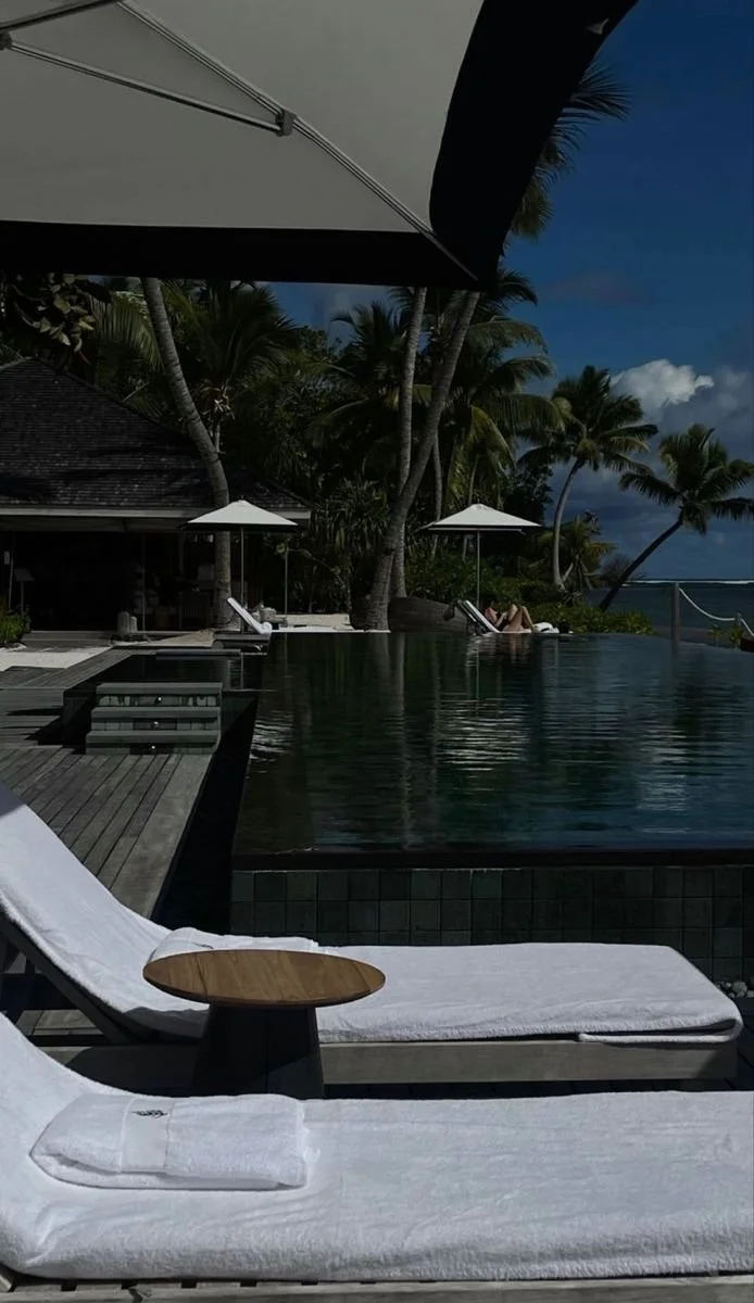 A poolside scene with white lounge chairs, a wooden side table, umbrellas, tropical palm trees, and a view of the ocean in the background under a partly cloudy sky.