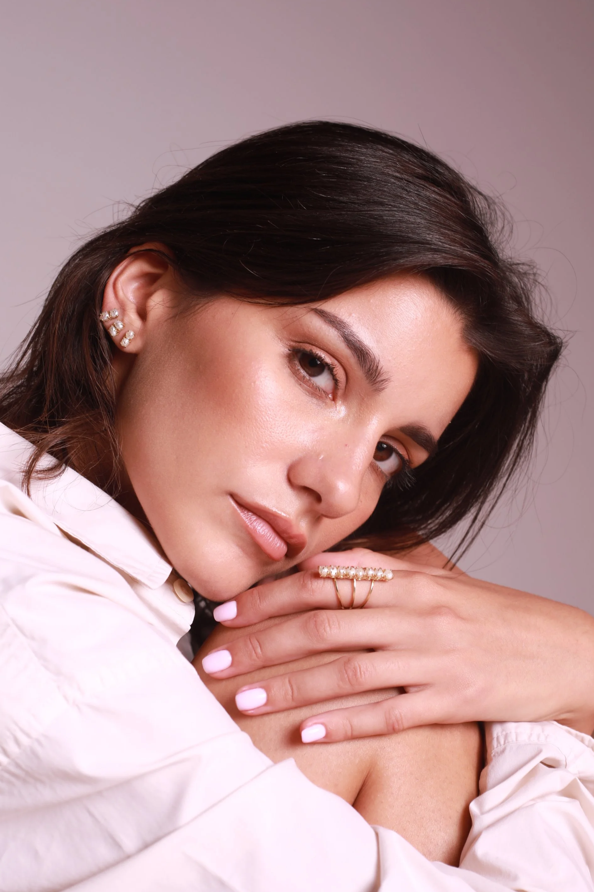 Jeune femme avec des boucles d'oreilles et une bague en perles, montrant un visage doux et regardant vers l'objectif, vêtue d'une chemise blanche, contre un fond gris neutre.
