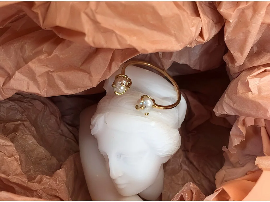 Une bague en or jaune avec des perles serties sur griffes repose sur la tete d'une sculpture. La tête est entourée de papier de soie rose poudré.