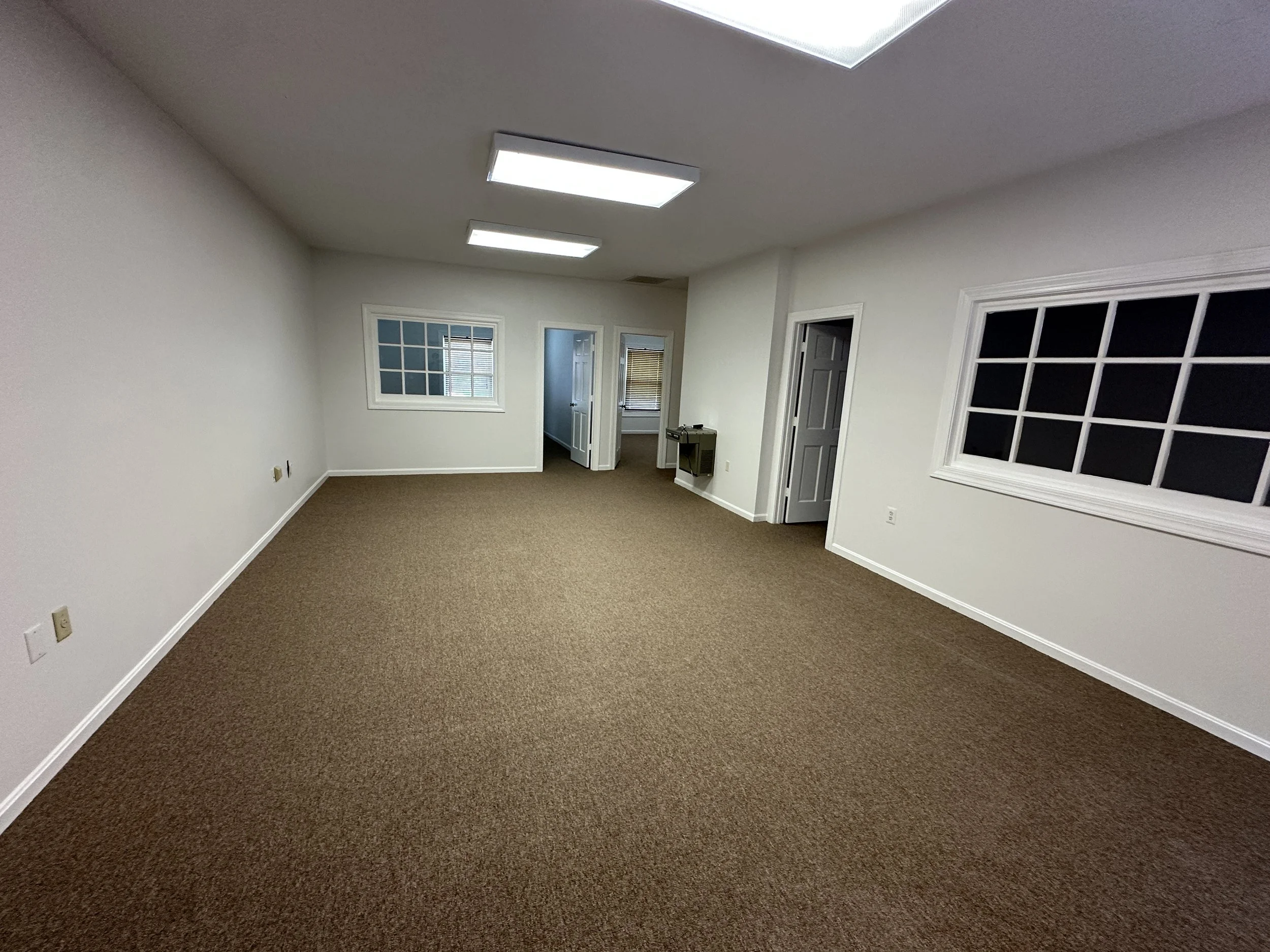 Empty room with beige carpet, white walls, ceiling with rectangular fluorescent light fixtures, several windows, and two open doors leading to other rooms.