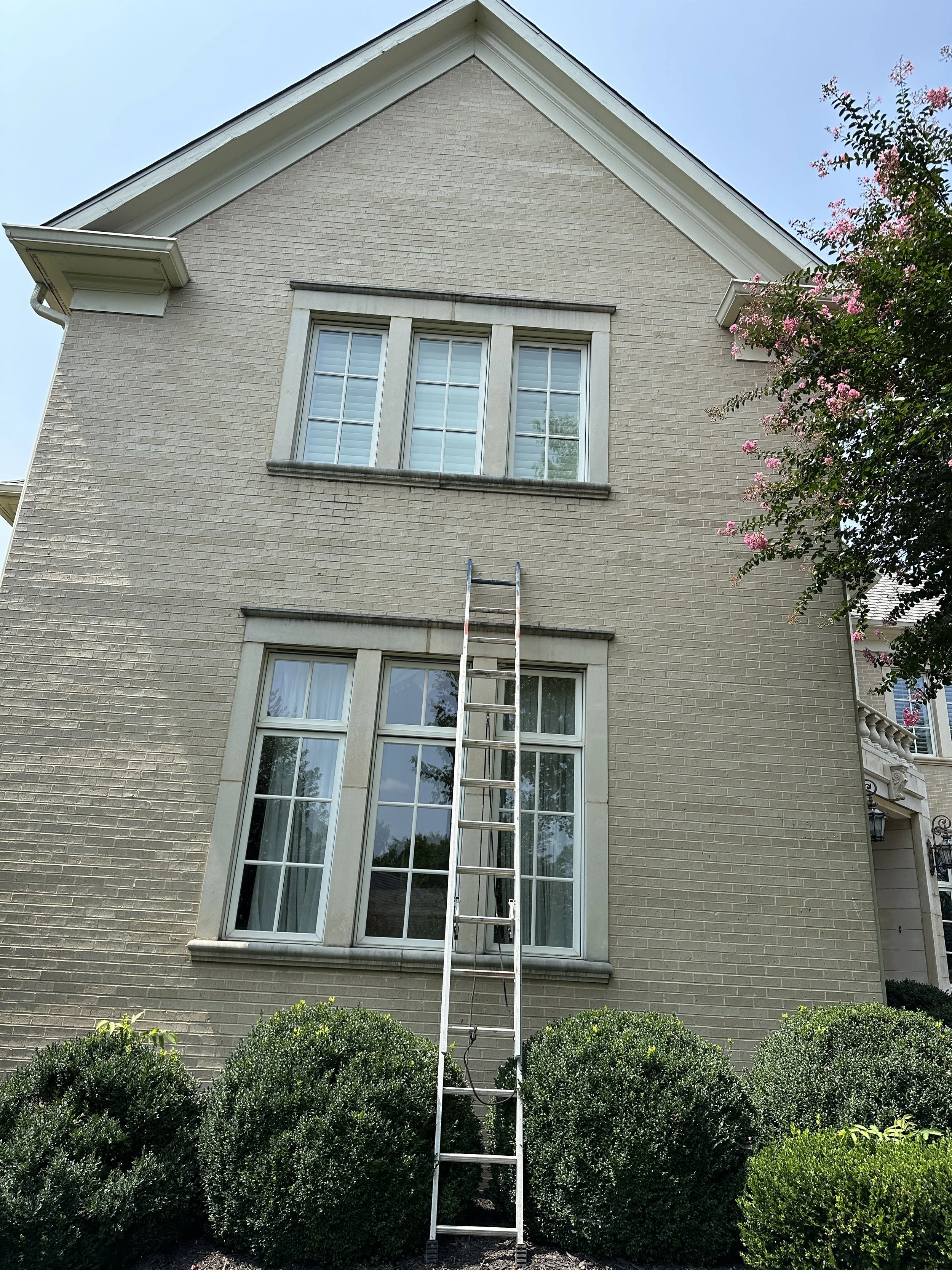 A beige brick house with a ladder leaning against the wall below two large windows on the first floor and three windows on the second floor. The house has a triangular roof and a small part of a pink flowering tree is visible on the right side of the