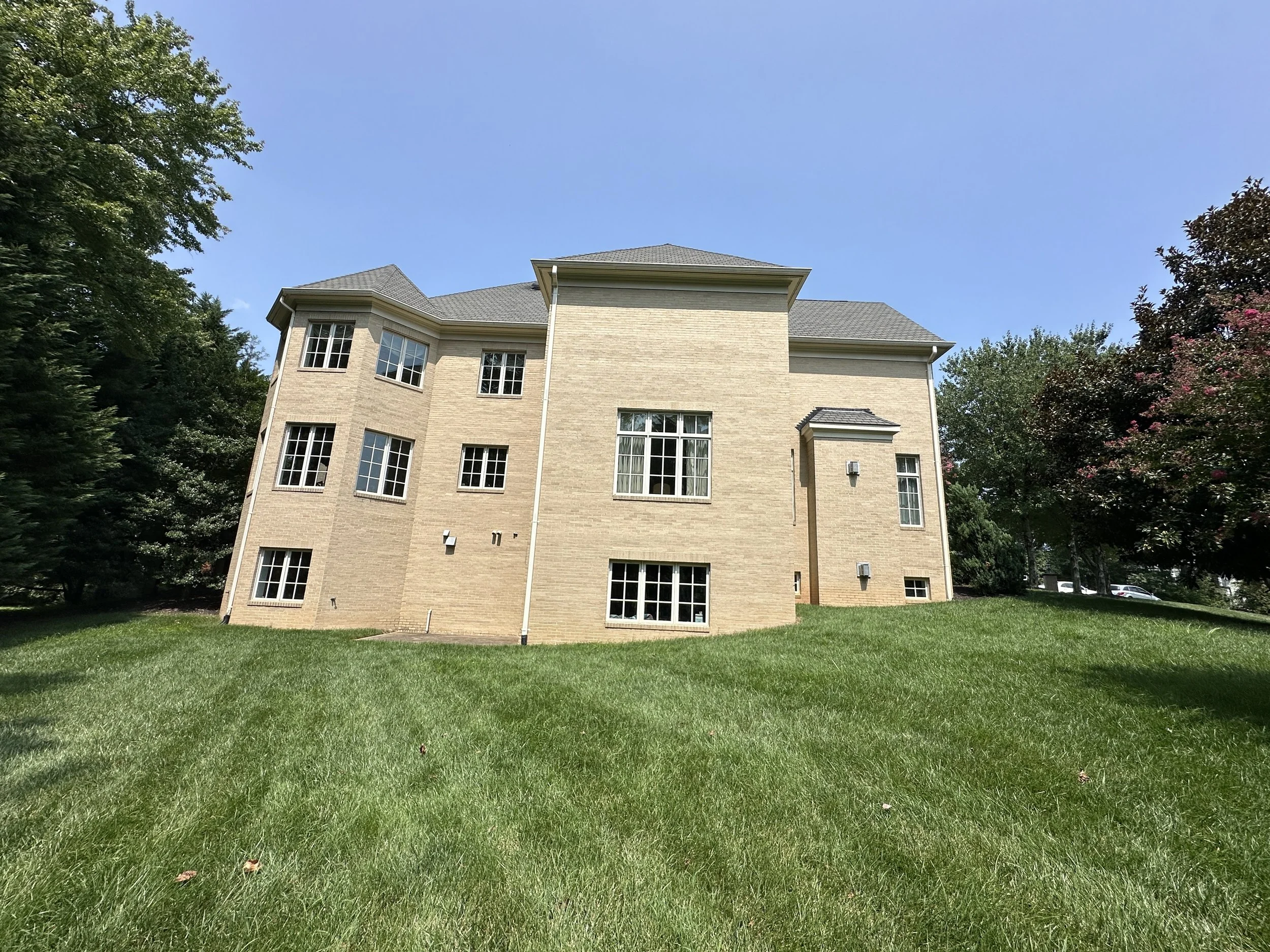 A multi-story beige brick house with a gray shingle roof, multiple large windows, and a well-kept green lawn.