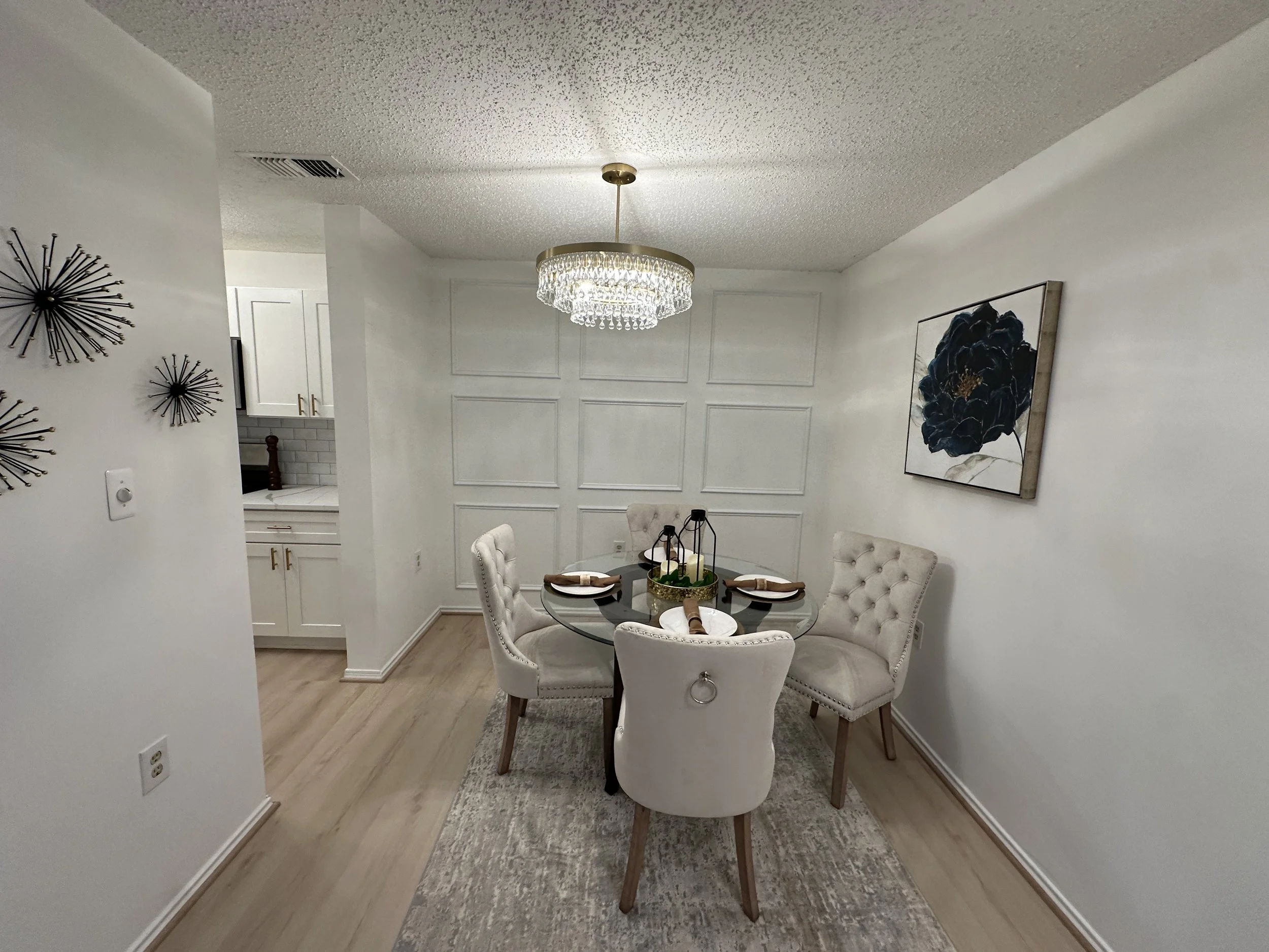 Dining room with a round glass table, four white tufted chairs, a chandelier, wall art, and a small gallery wall with metal starburst designs.