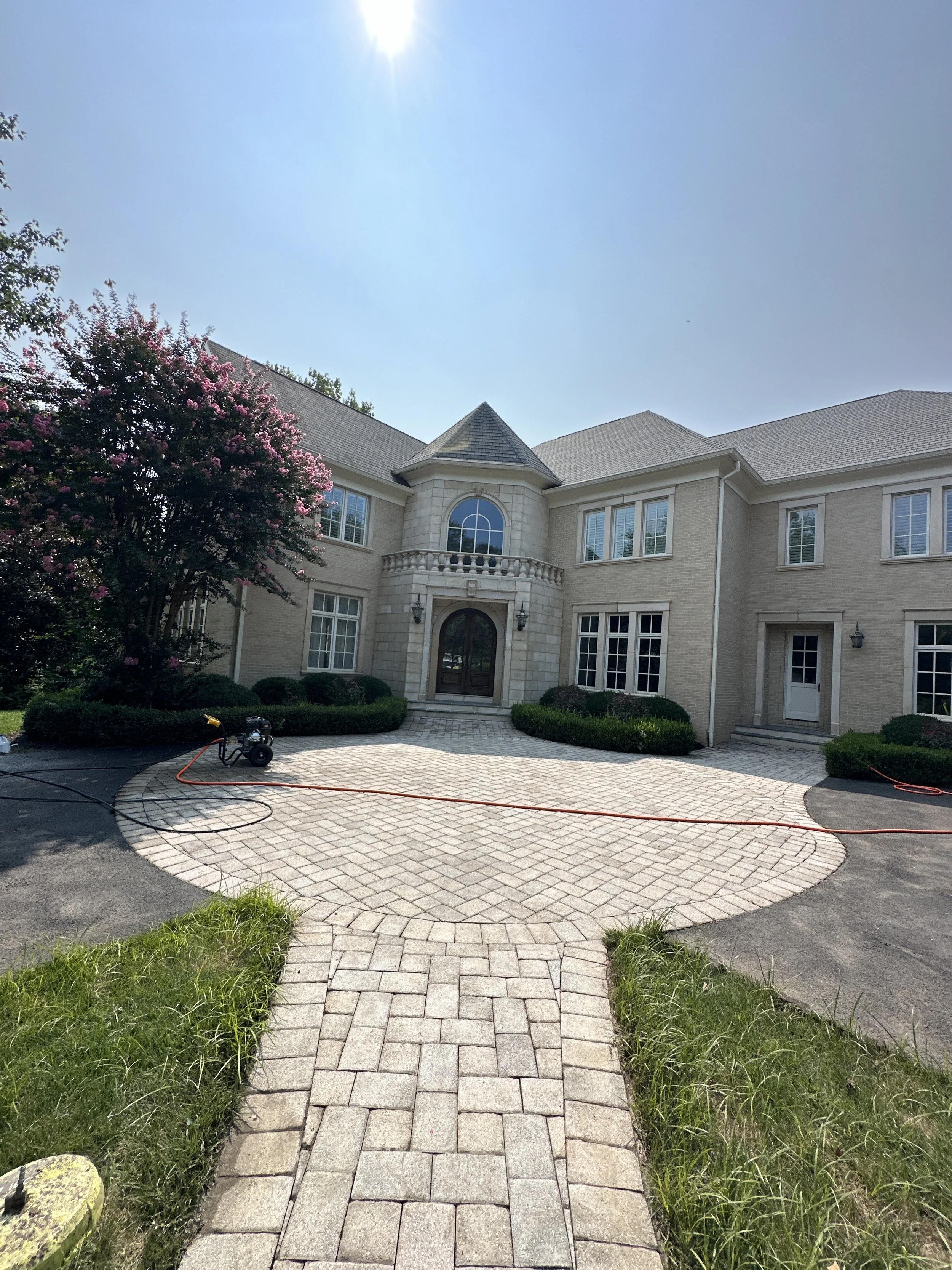 View of a large beige brick house with a curved driveway, a pathway leading to the front door, lush shrubs, trees, and a flowering tree on the left, under a clear sunny sky.