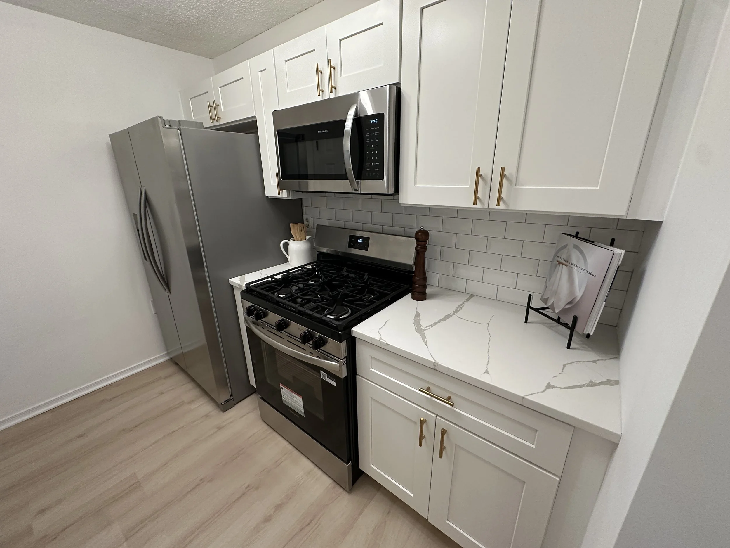 Kitchen with white cabinets, a stainless steel refrigerator, a microwave, and a stove. The countertops are white with gray veining, and there's a small cookbook stand on the right side.