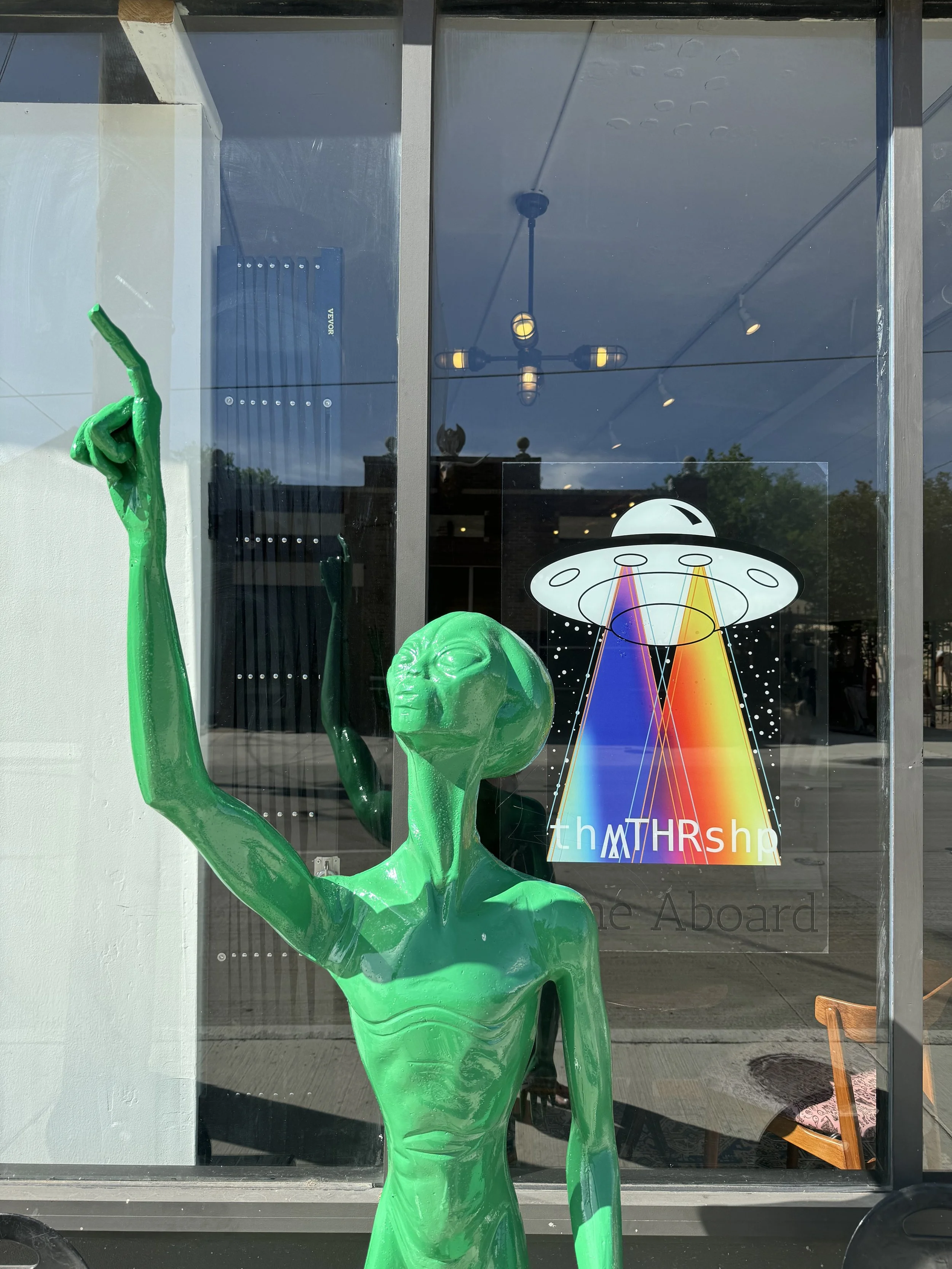 Green alien sculpture with an elongated neck and head, standing inside a building and facing out through a glass window. The alien appears to be raising one arm with a finger pointing upward. Behind the alien, there's a window with a colorful graphic of a UFO emitting rainbow beams, and the words 'the THRship' and 'Aboard' visible on the window.