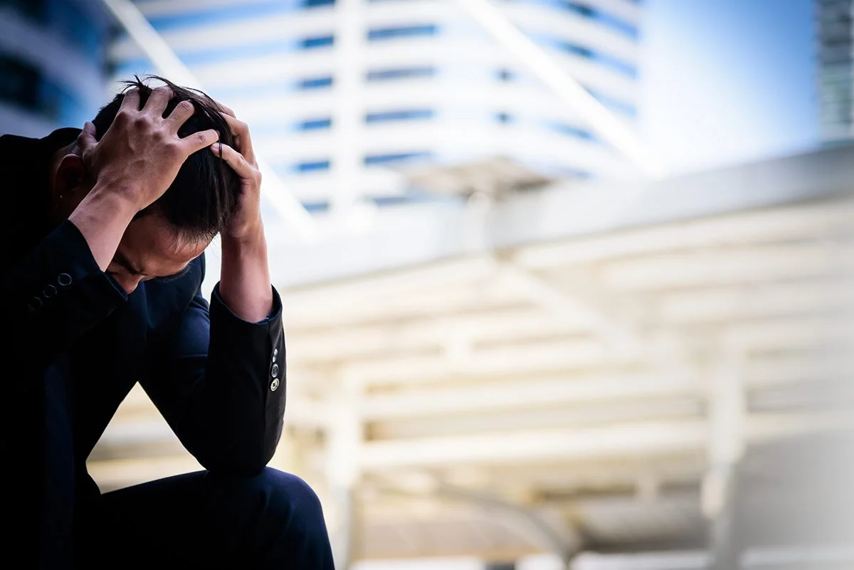 A man in a black suit sitting outdoors with his hands on his head, looking distressed.