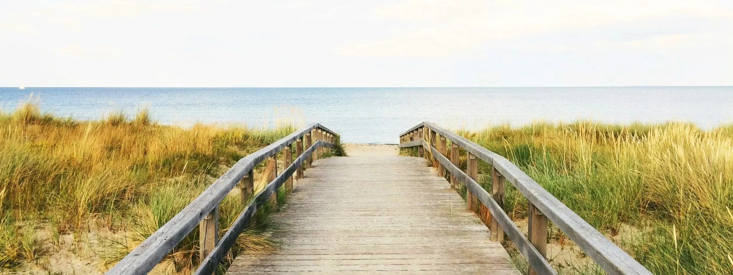 Wooden walkway leading to a beach with sand dunes and tall grass, overlooking the calm ocean under a partly cloudy sky.