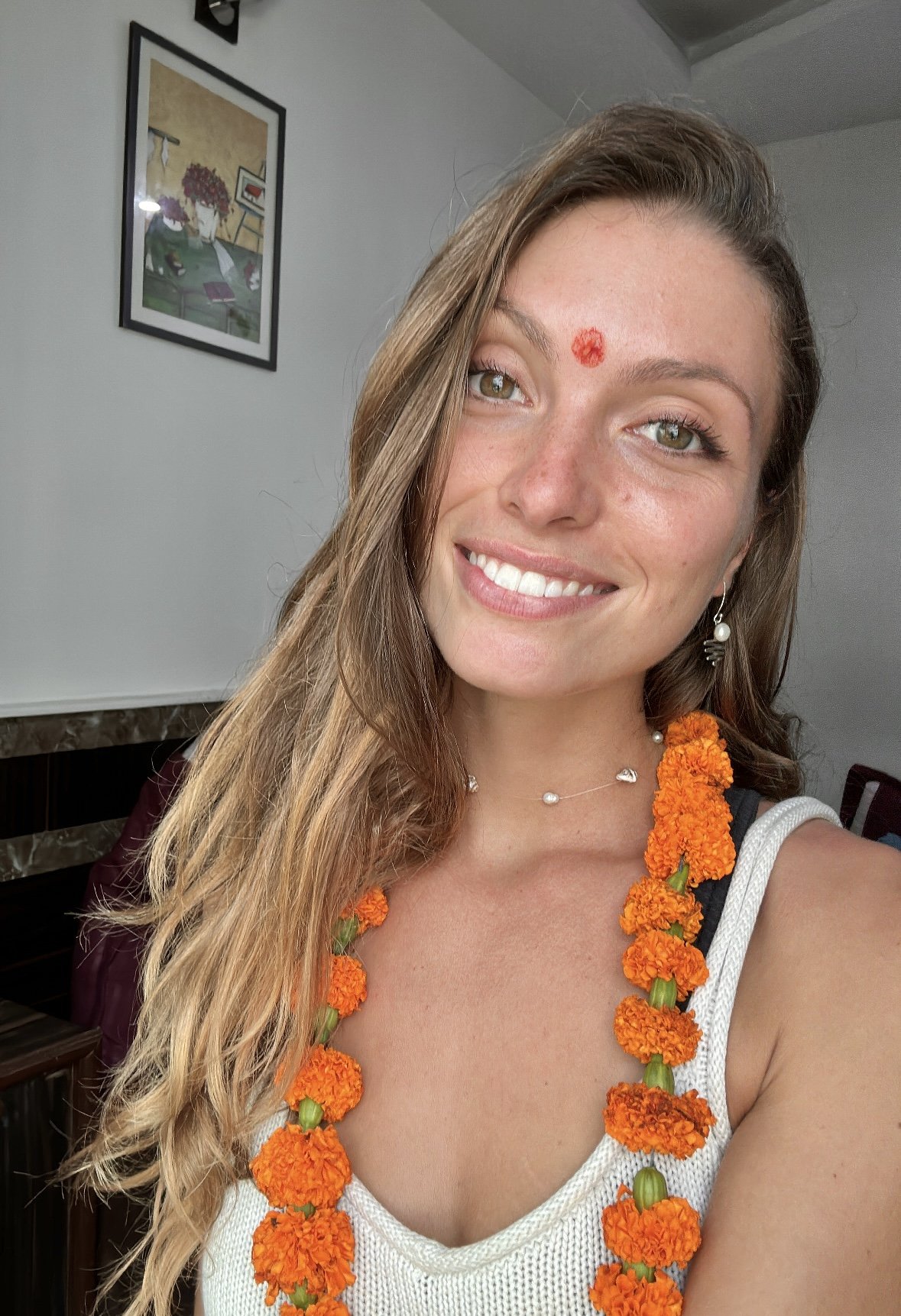 A young woman with light brown hair, smiling, wearing a white sleeveless top, a necklace with small pearls, earrings, and an orange flower garland around her neck, indoors with a framed picture on the wall in the background.
