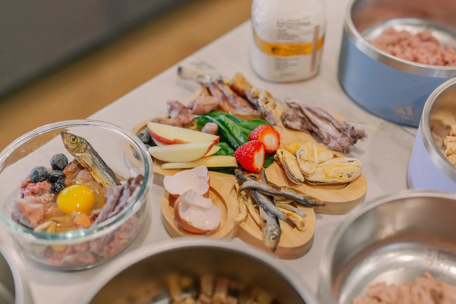 A wooden tray with strawberries, apple slices, and dried fish on a table next to bowls of pet food, some with fish and eggs, and a can of pet food in the background.