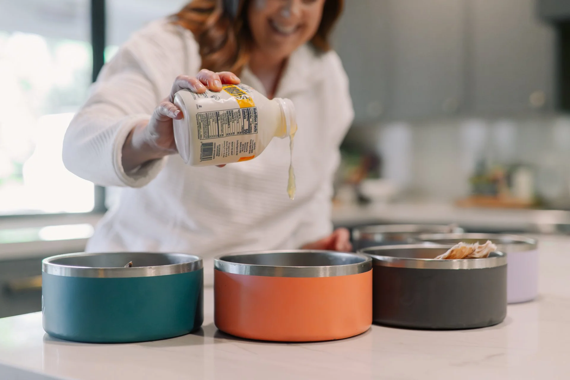 A woman in a white shirt is pouring a creamy liquid into colorful containers on a kitchen counter.
