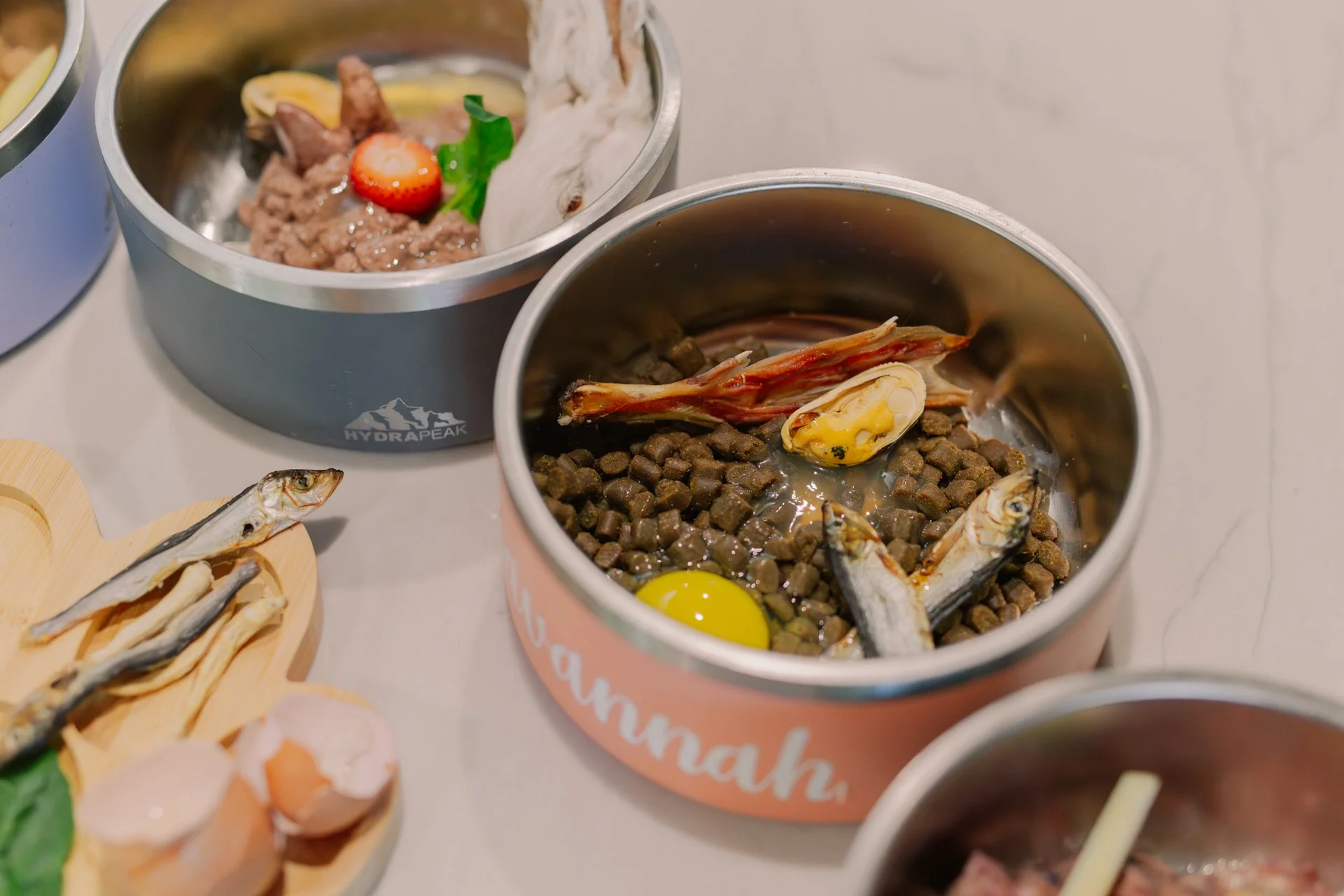 Open cans of pet food containing fish pieces, meat, and other ingredients, along with a raw egg yolk in one of the cans, on a countertop.