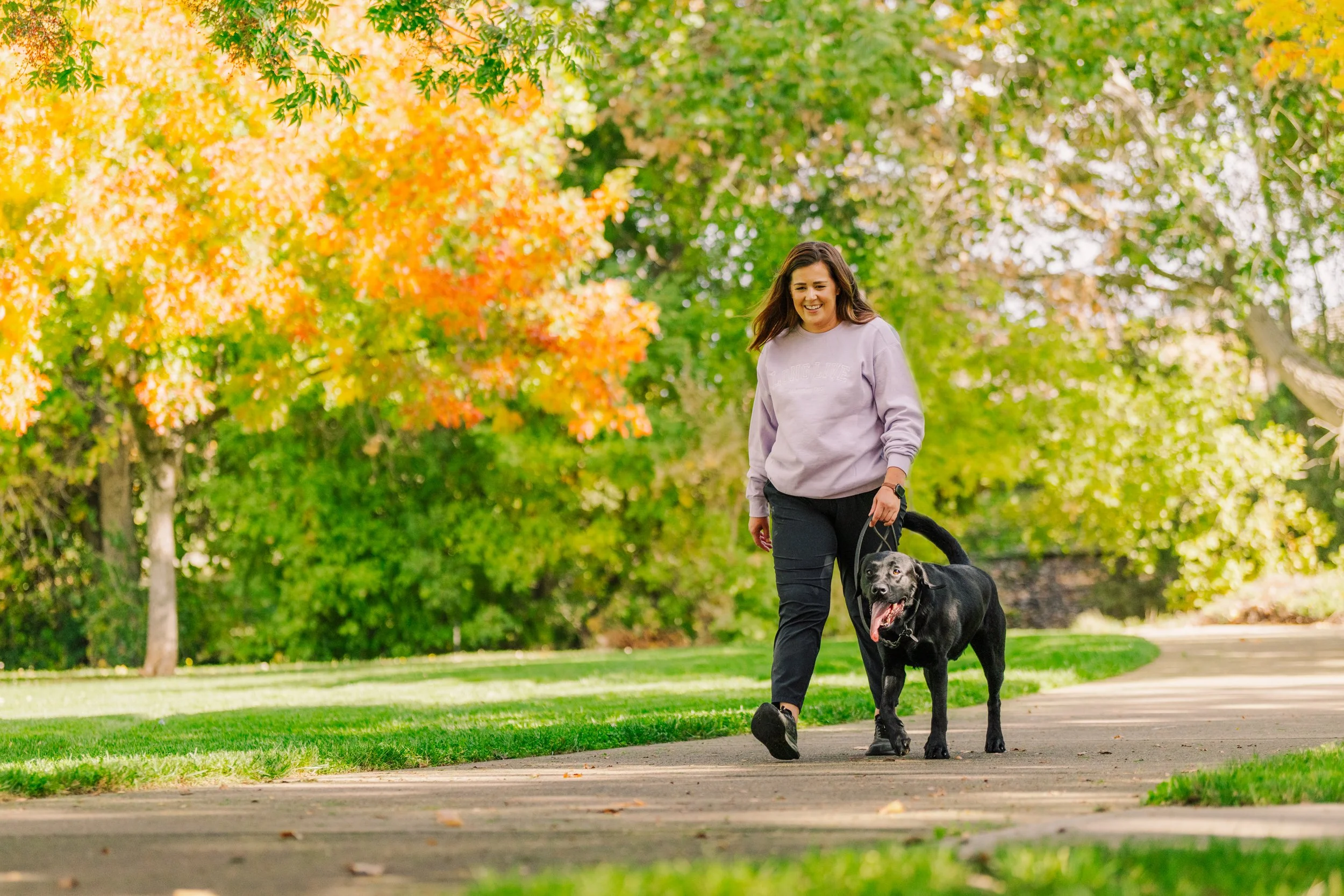 A woman walking her black dog along a park path with fall foliage in the background.