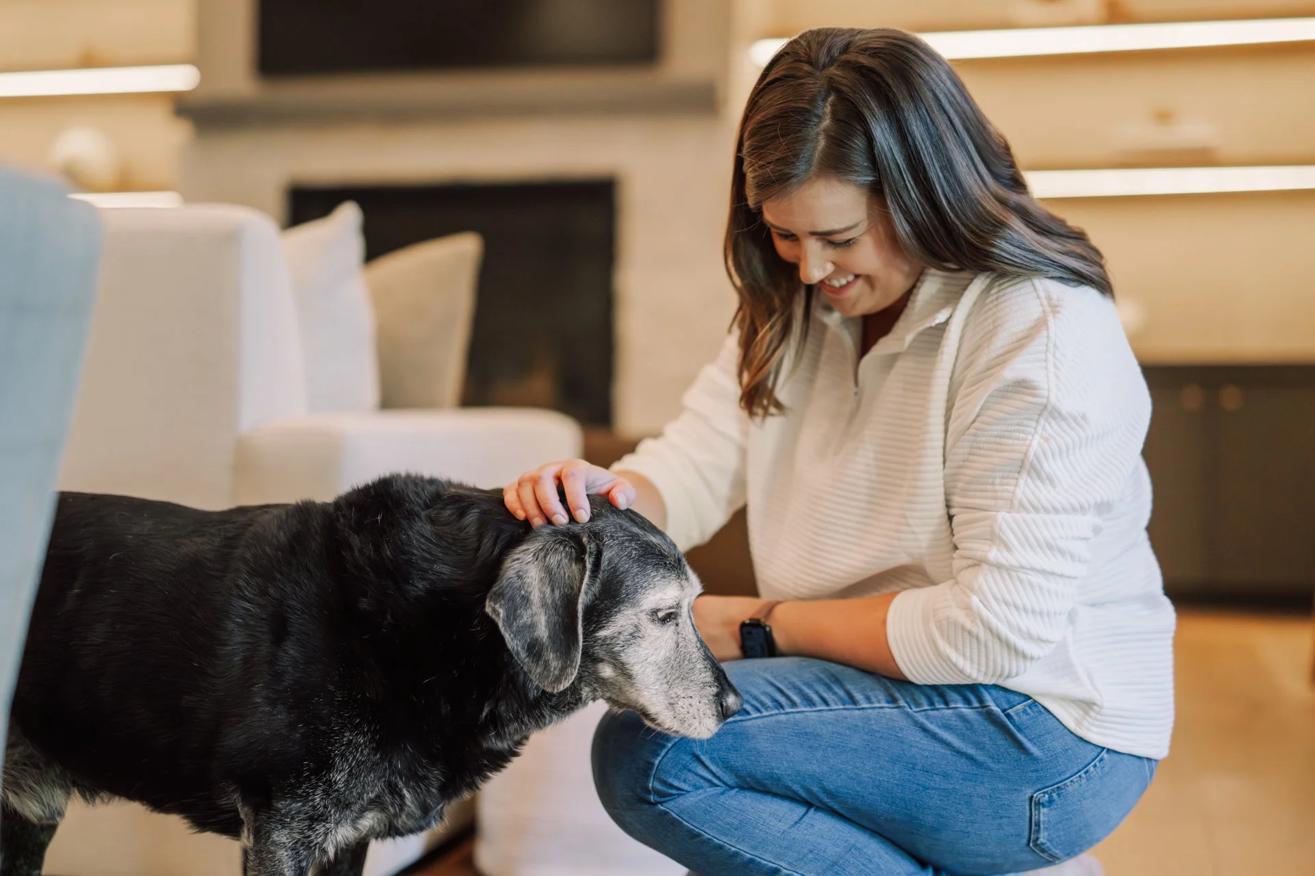 A woman in a beige sweater and blue jeans kneels on the floor, smiling as she gently pets a large black and gray dog lying on the floor.