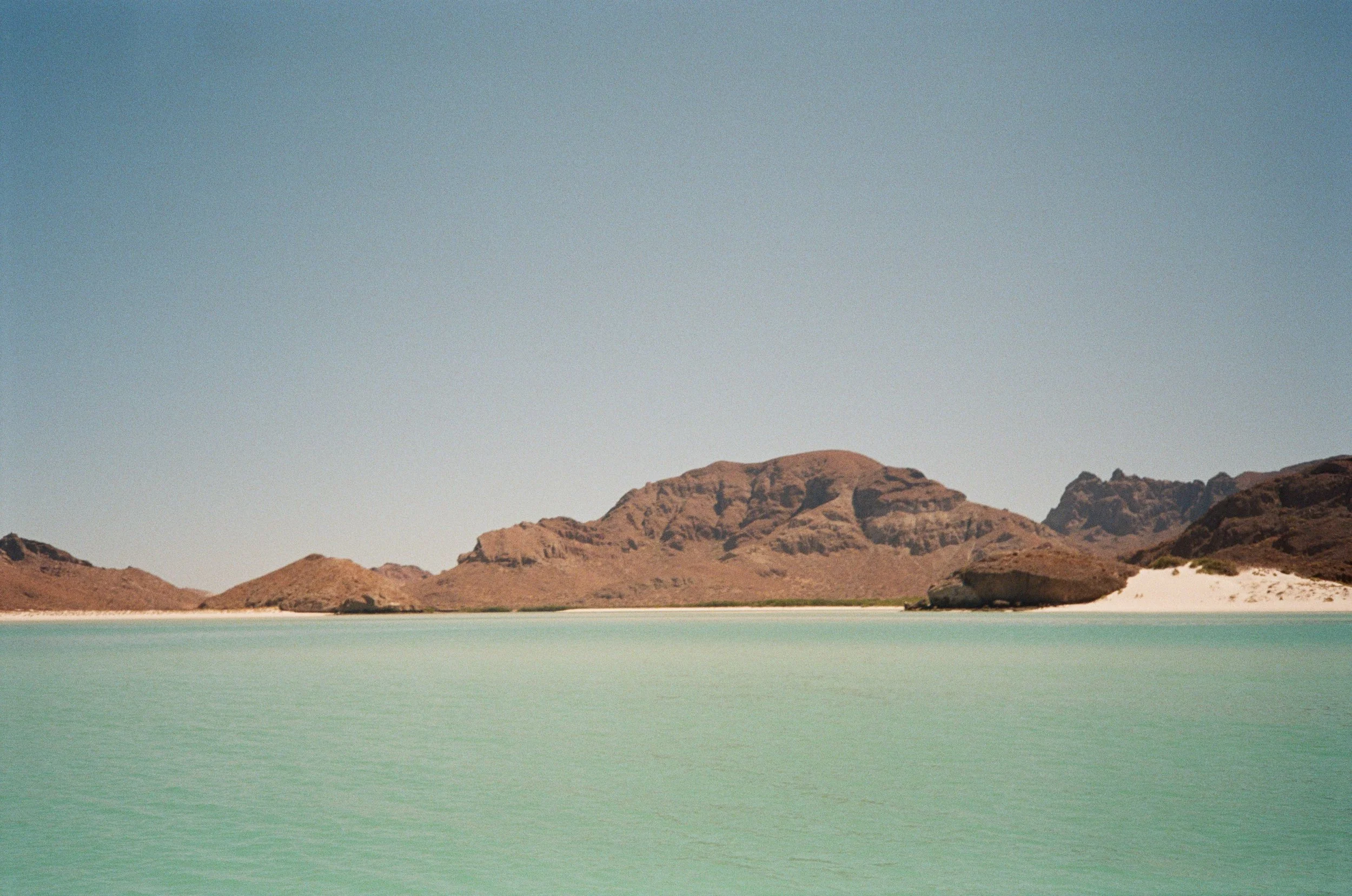 A body of turquoise water with brown mountains and clear blue sky in the background.